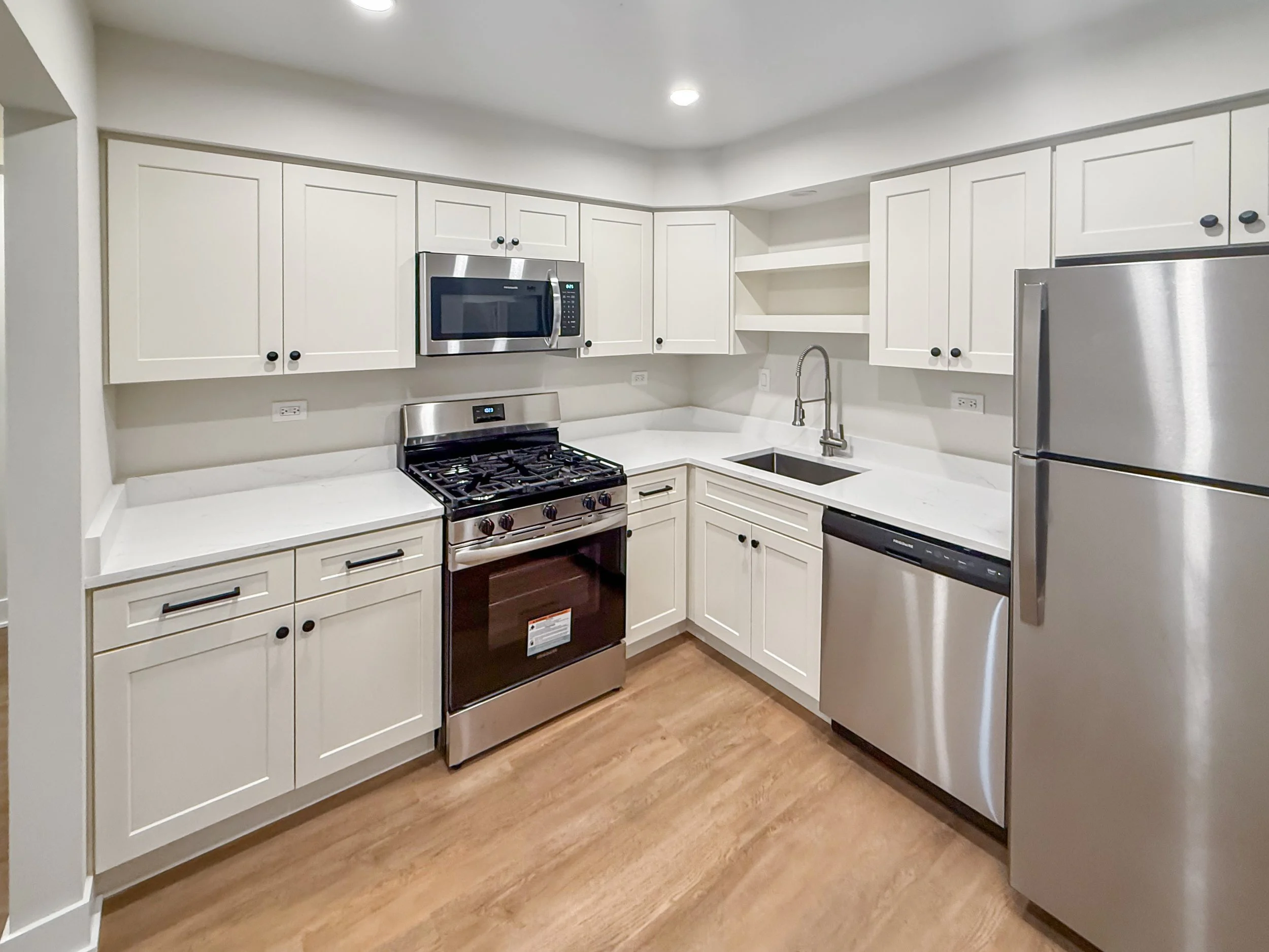 Modern kitchen with white cabinets, stainless steel appliances, a black stovetop, microwave, dishwasher, and a refrigerator with wood flooring.