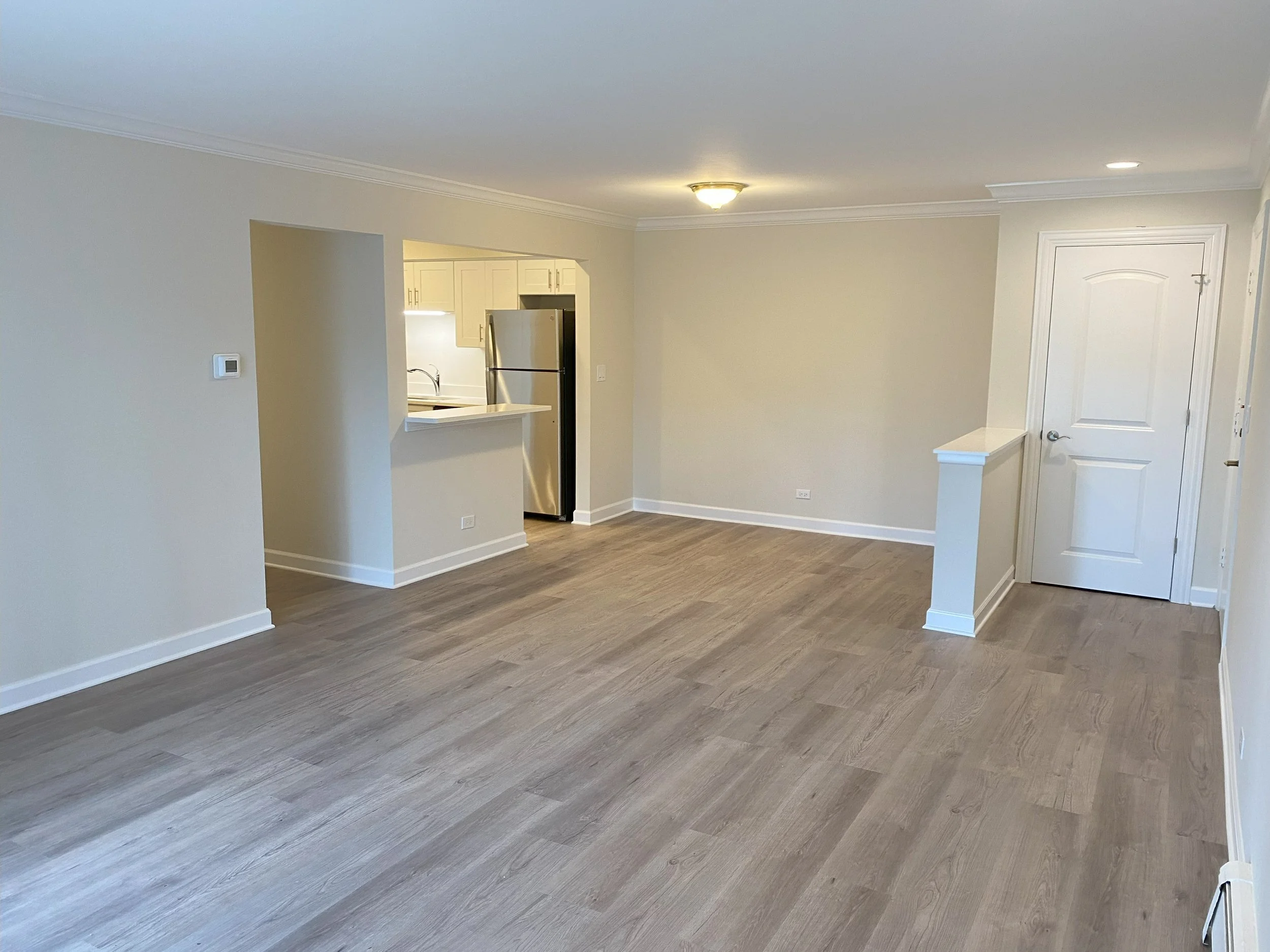 Empty living room with beige walls, wood flooring, and an open kitchen in the background with stainless steel appliances.