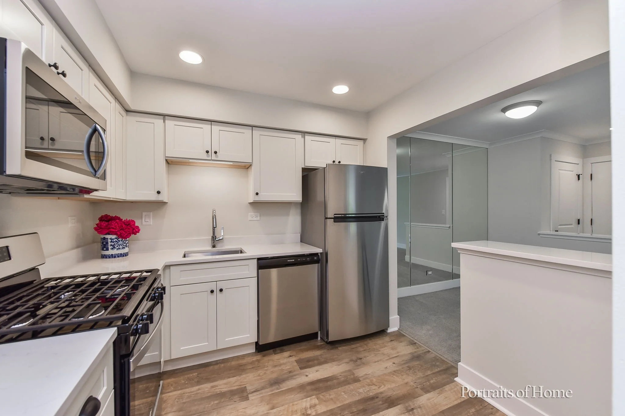 Kitchen with white cabinets, stainless steel refrigerator, microwave, and dishwasher. There is a pot with pink flowers on the counter, a sink with a faucet, and wood flooring. A pass-through window opens to another room with a glass-enclosed area.