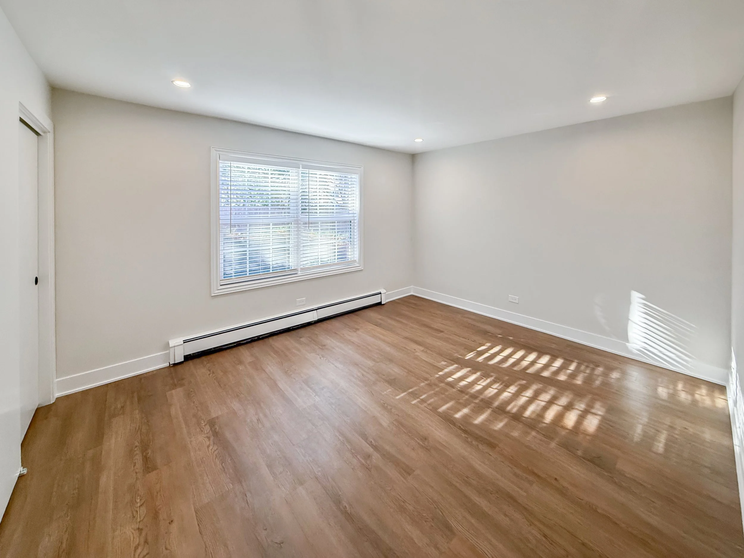 Empty room with white walls, hardwood floors, window with blinds, and baseboard heater.