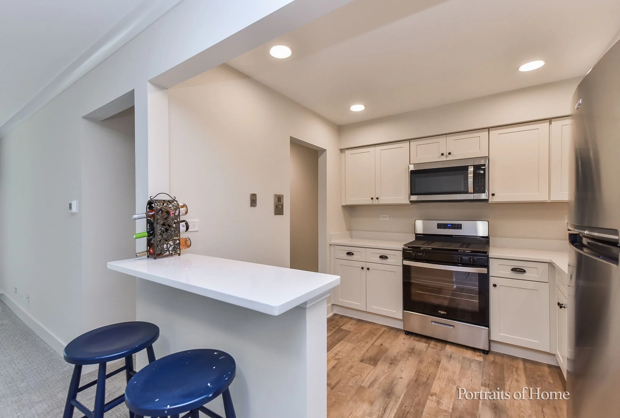 Modern kitchen with white cabinets, stainless steel appliances including a microwave and oven, wooden floor, and a small breakfast bar with two blue stools.
