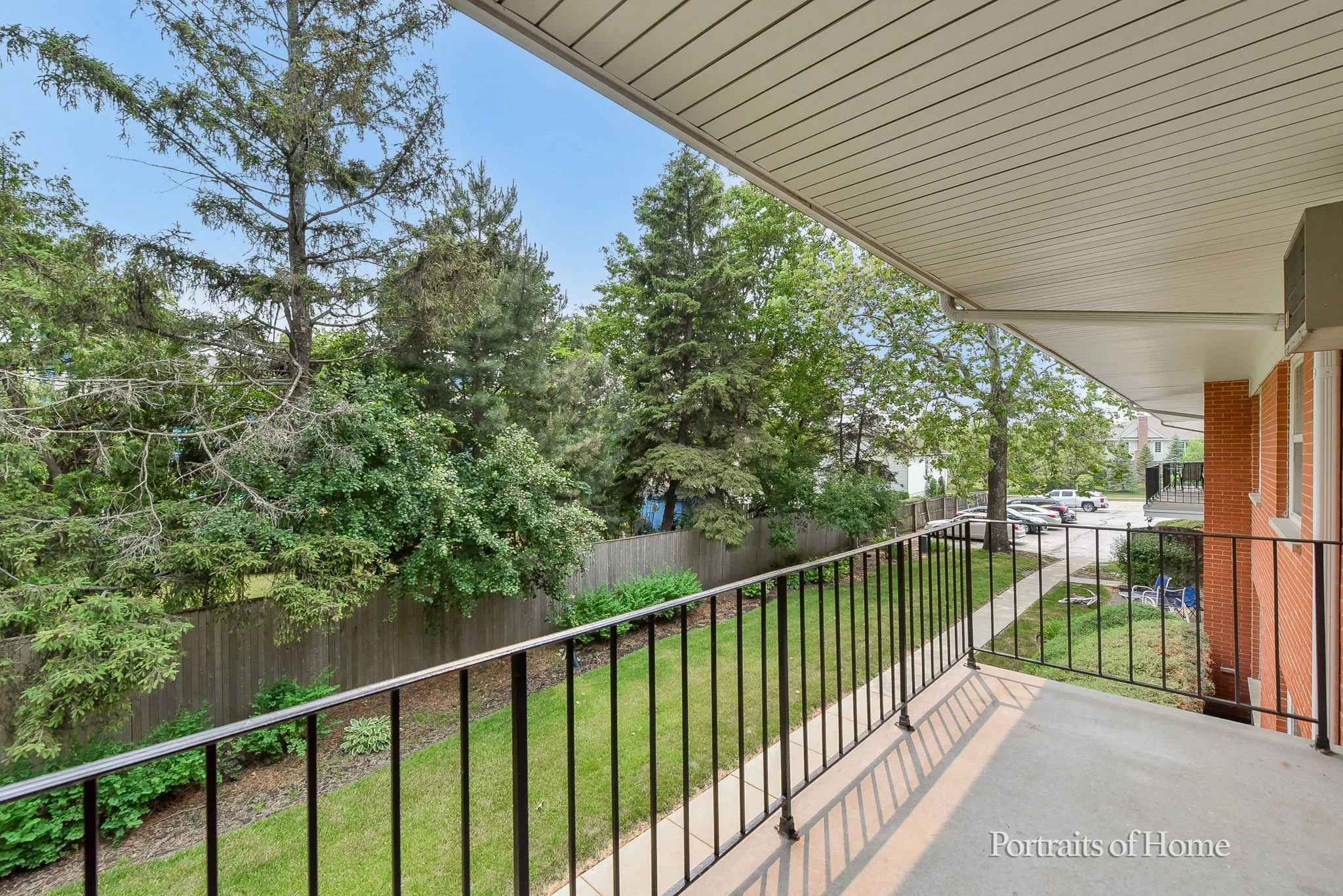 Balcony with black metal railing overlooking a green backyard with trees and a parking lot in the distance.