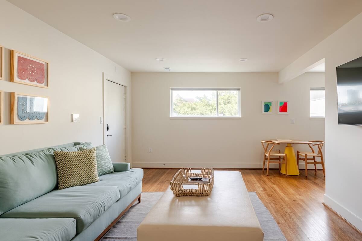 Bright living room with a light green sofa, a wooden coffee table, wall art, windows, and a small yellow table with chairs.