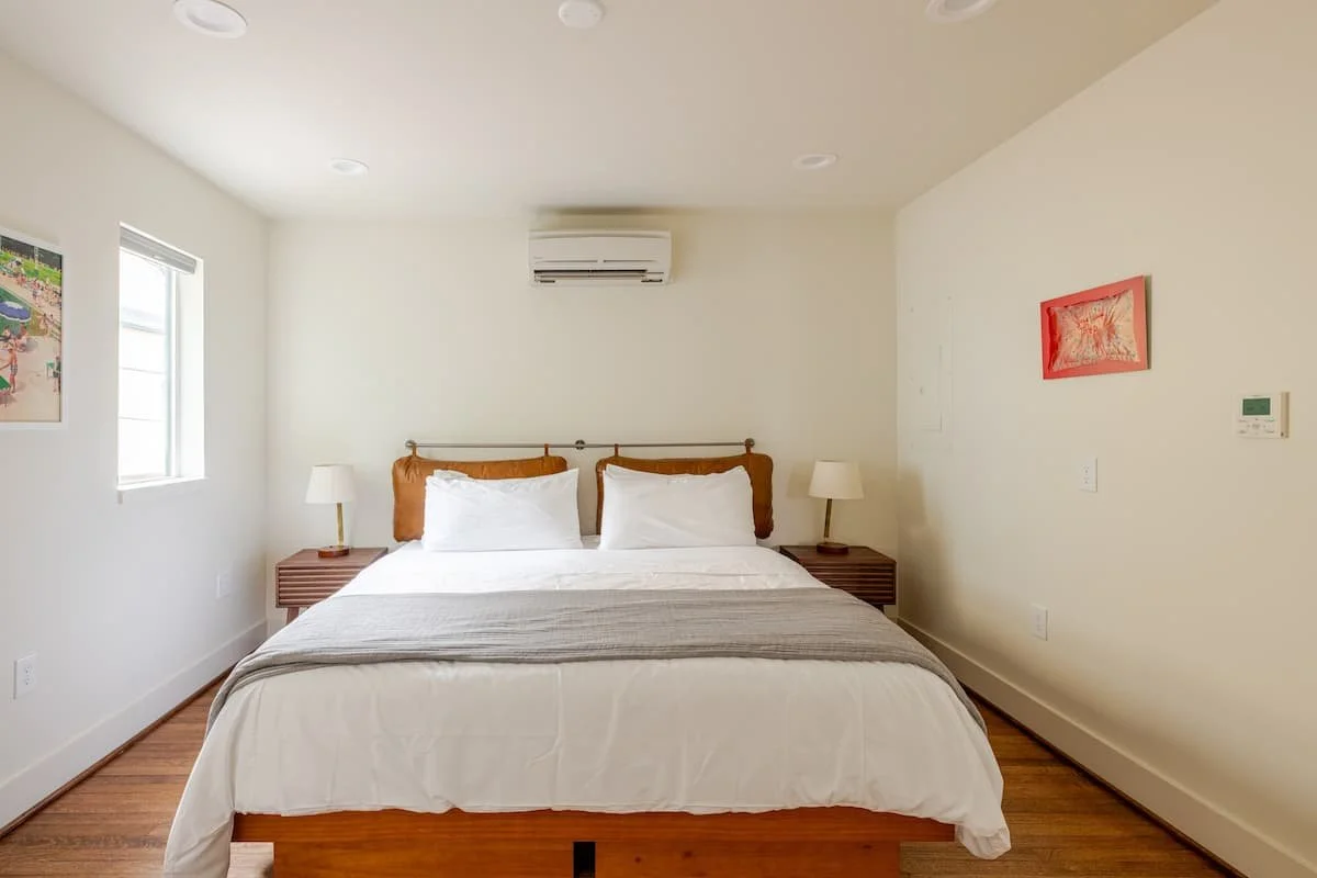 A neatly made bed with white bedding and two pillows in a bedroom with white walls, two side tables with lamps, solar panel on the wall, and artwork on the walls.