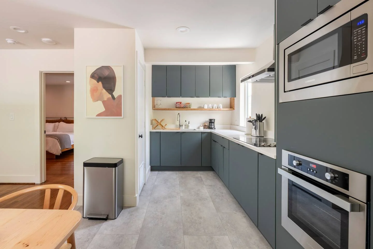 Modern kitchen with gray cabinets, a built-in microwave, oven, and a sink beneath a window. It features a wooden shelf with cups and a coffee maker, and a glimpse of a bedroom through an open door.