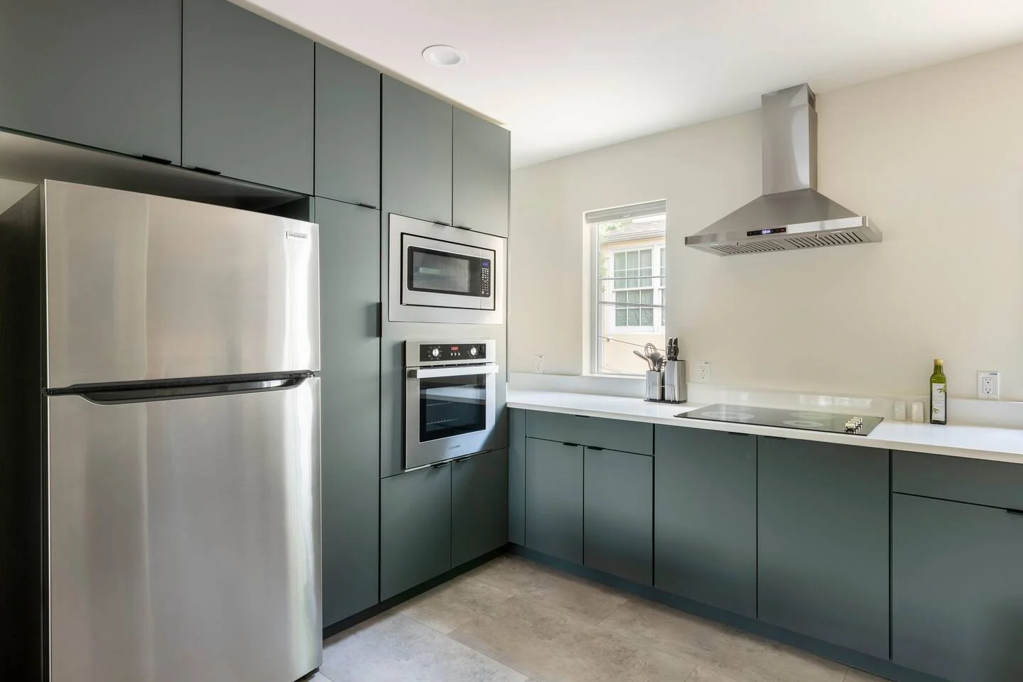 Modern kitchen with gray cabinets, stainless steel refrigerator, built-in microwave and oven, window, white countertop, stove, range hood, and door handle.