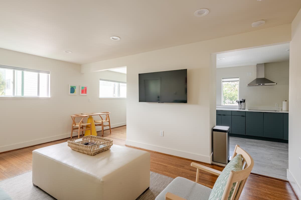 Living room with white walls, hardwood floors, a wall-mounted TV, a beige ottoman with a tray, a chair with patterned cushion, and a small dining area with a table and chairs, adjacent to a kitchen with dark cabinets and a window.