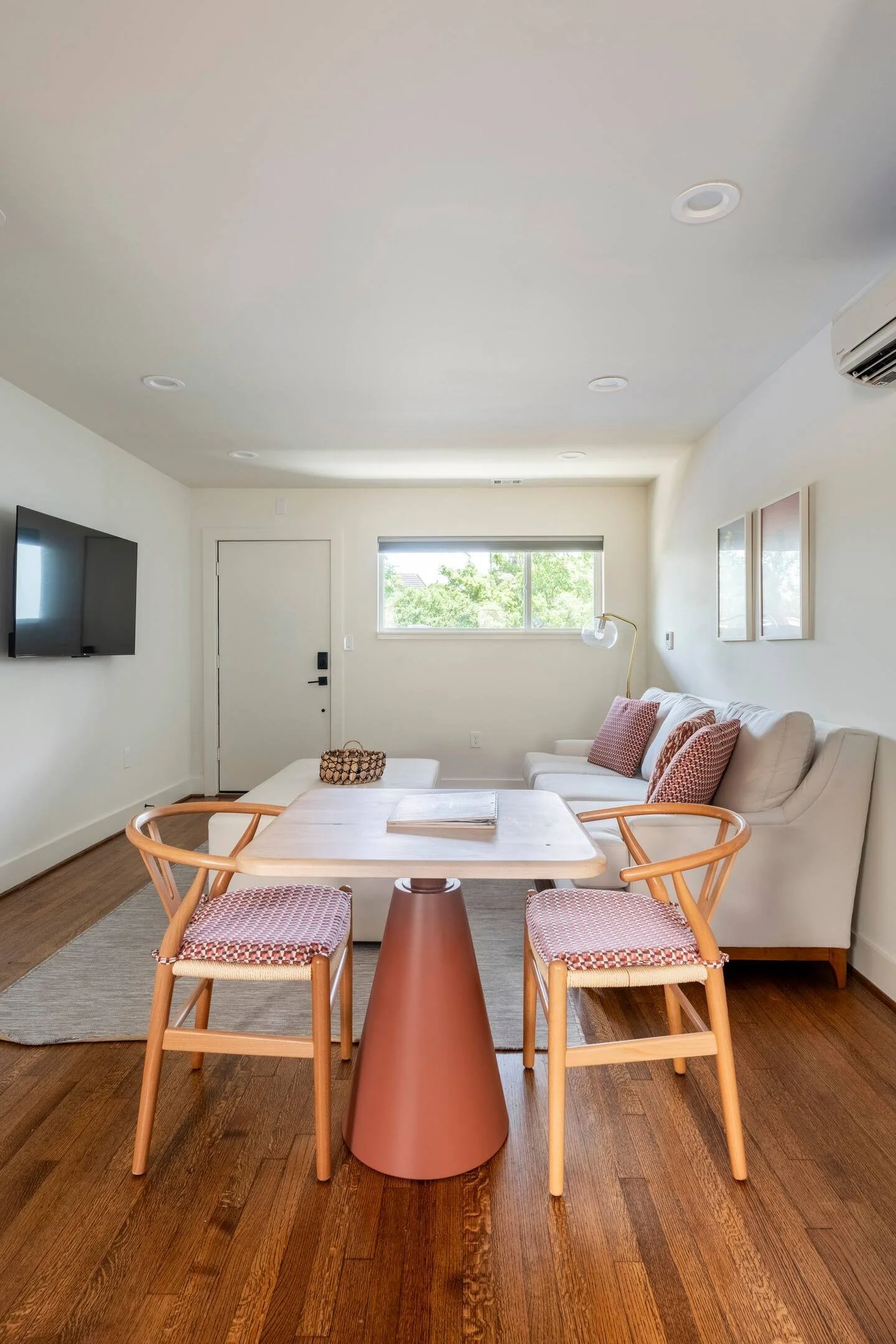 A cozy living room with a white sofa, pink accent pillows, a wooden dining table with four chairs, a wall-mounted TV, a window with a view of greenery, and wooden flooring.