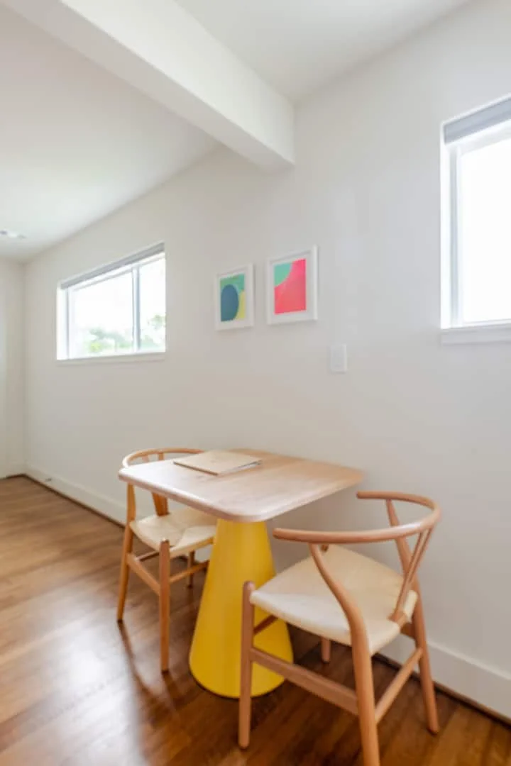A small wooden table with a yellow base, two wooden chairs with white cushions, two colorful abstract art prints on a white wall, and two windows providing natural light in a minimalistic room.