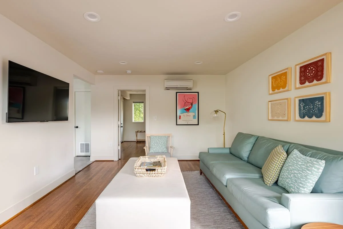 Living room with a teal sofa, patterned throw pillows, a white ottoman, wall art, a wall-mounted TV, a floor lamp, and a doorway leading to another room with hardwood floors.