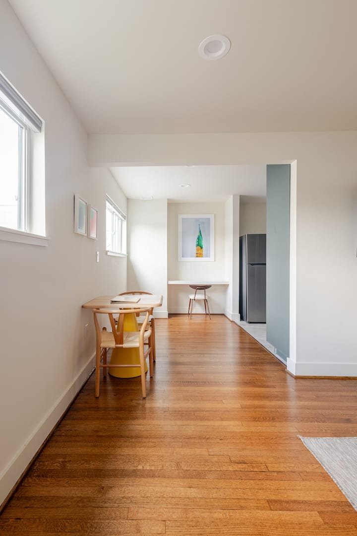 A minimalist dining area with a wooden table and chairs, a small counter space with a stool, and a refrigerator in the background, with white walls and hardwood flooring.