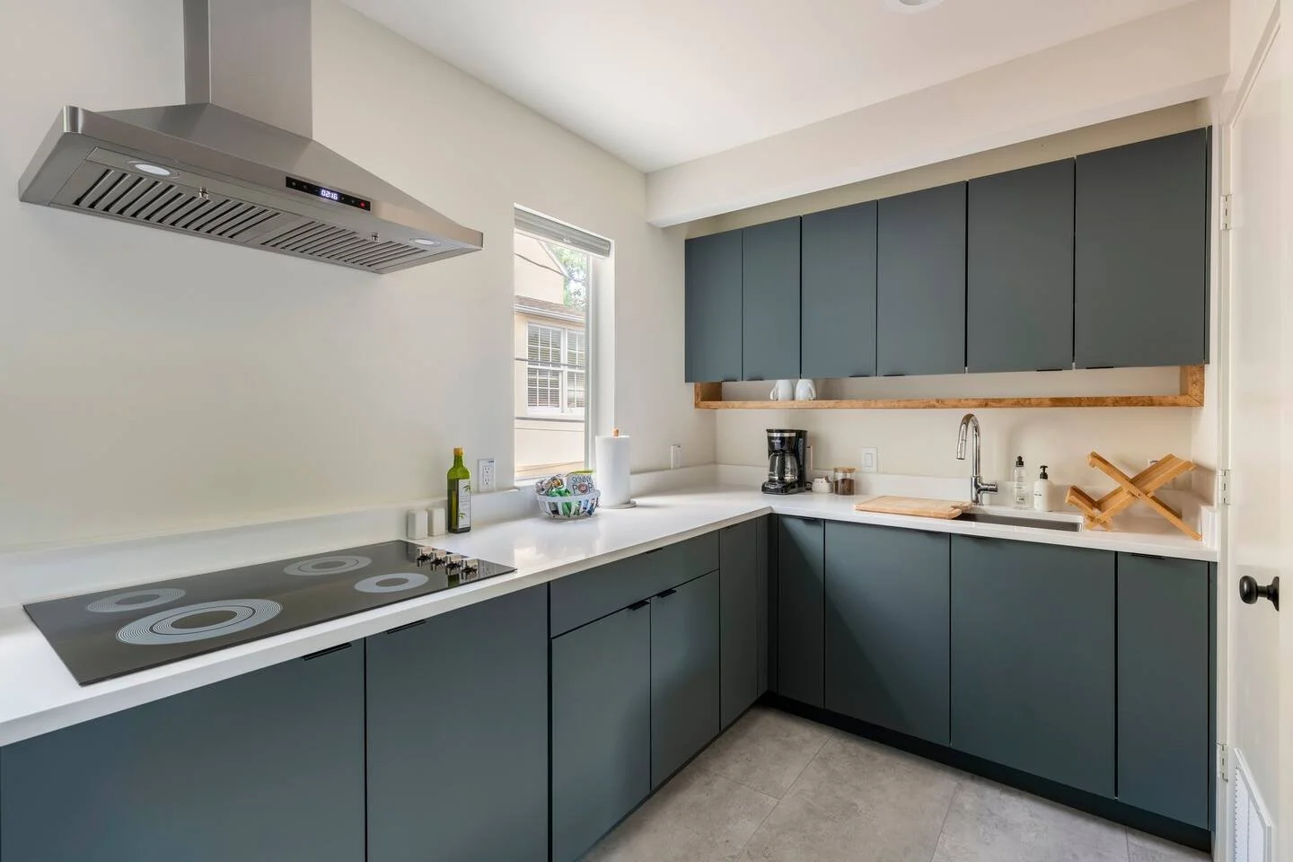 Modern kitchen with dark blue cabinets, white countertop, stainless steel range hood, stovetop, small window, coffee maker, dish soap, and a paper towel holder.