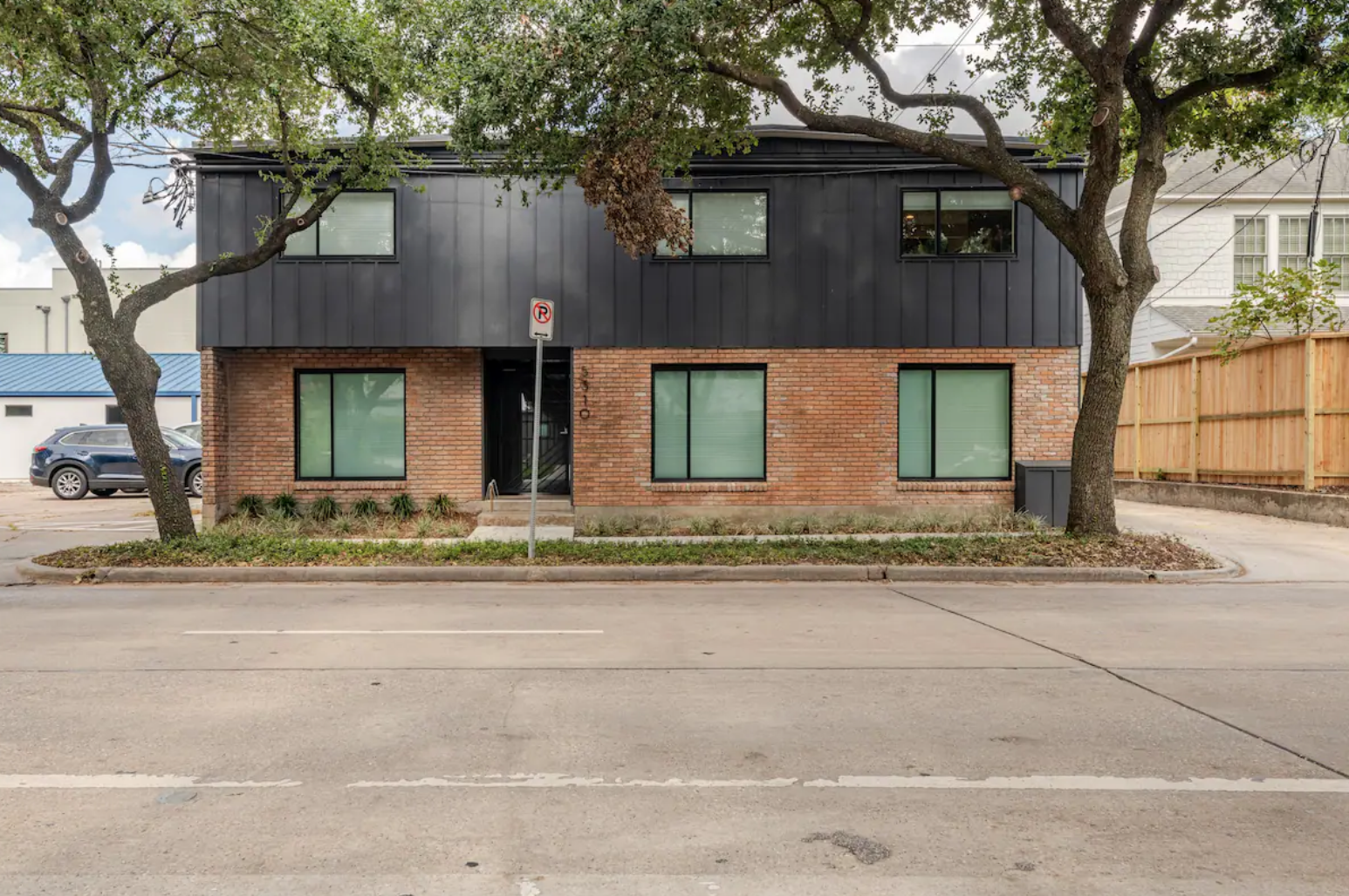 A modern two-story building with a brick lower floor and black upper floor, surrounded by trees and a wooden fence, on a city street.