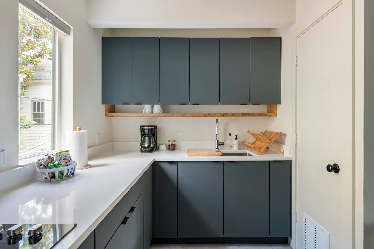 Modern kitchen with blue cabinets, white countertops, window, coffee maker, paper towel roll, dish drying rack, sink, soap bottles, and decorative vases.