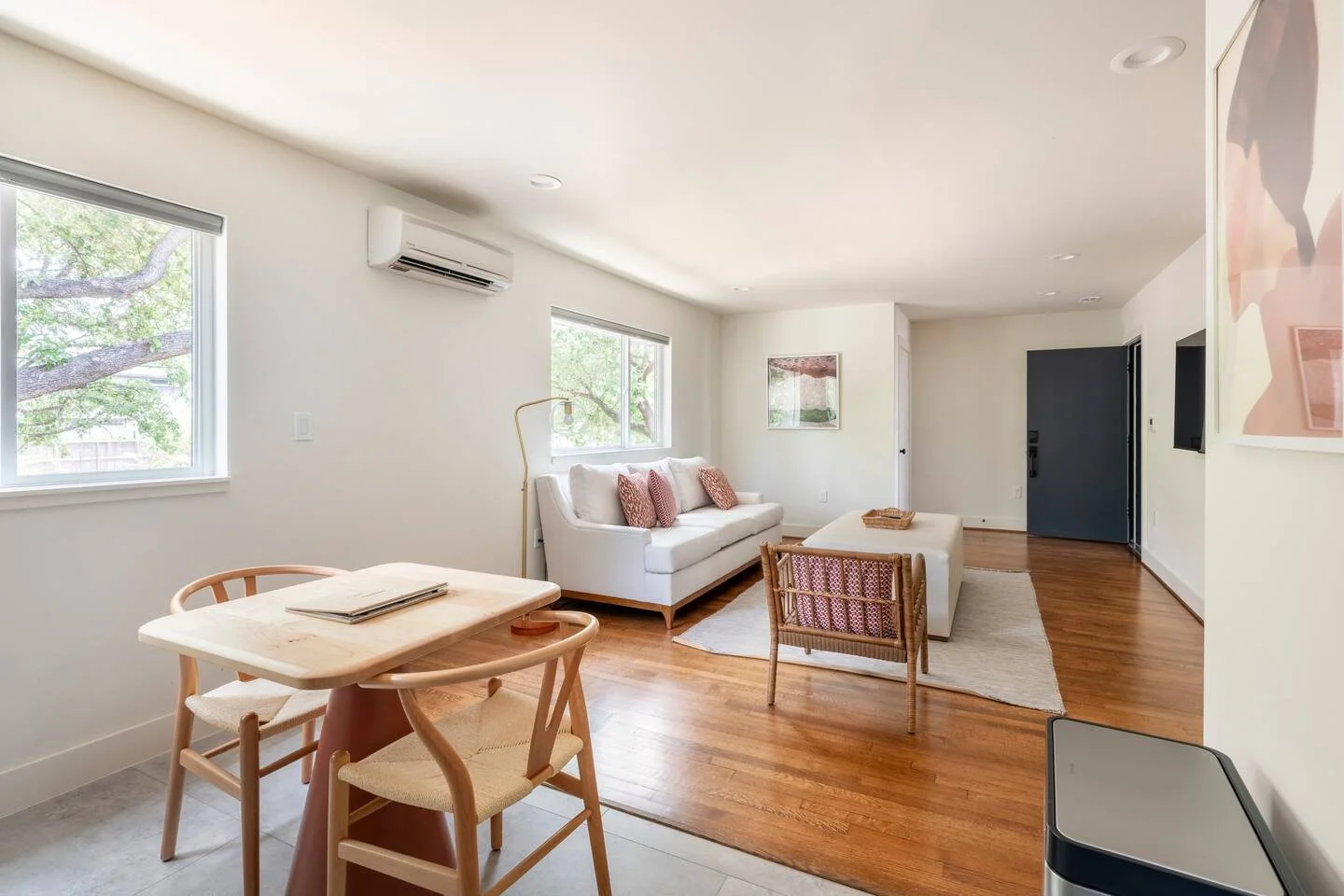 Bright living room with white walls, a white sofa with pink pillows, a small wooden chair, a rectangular ottoman, and a beige area rug. Two windows show tree branches outside. Wooden flooring extends throughout the space.