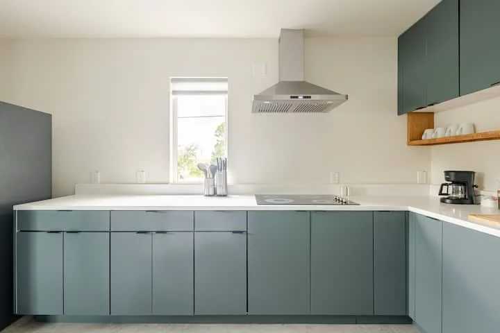 Modern kitchen with blue cabinets, white countertop, stainless steel range hood, window, and coffee maker.