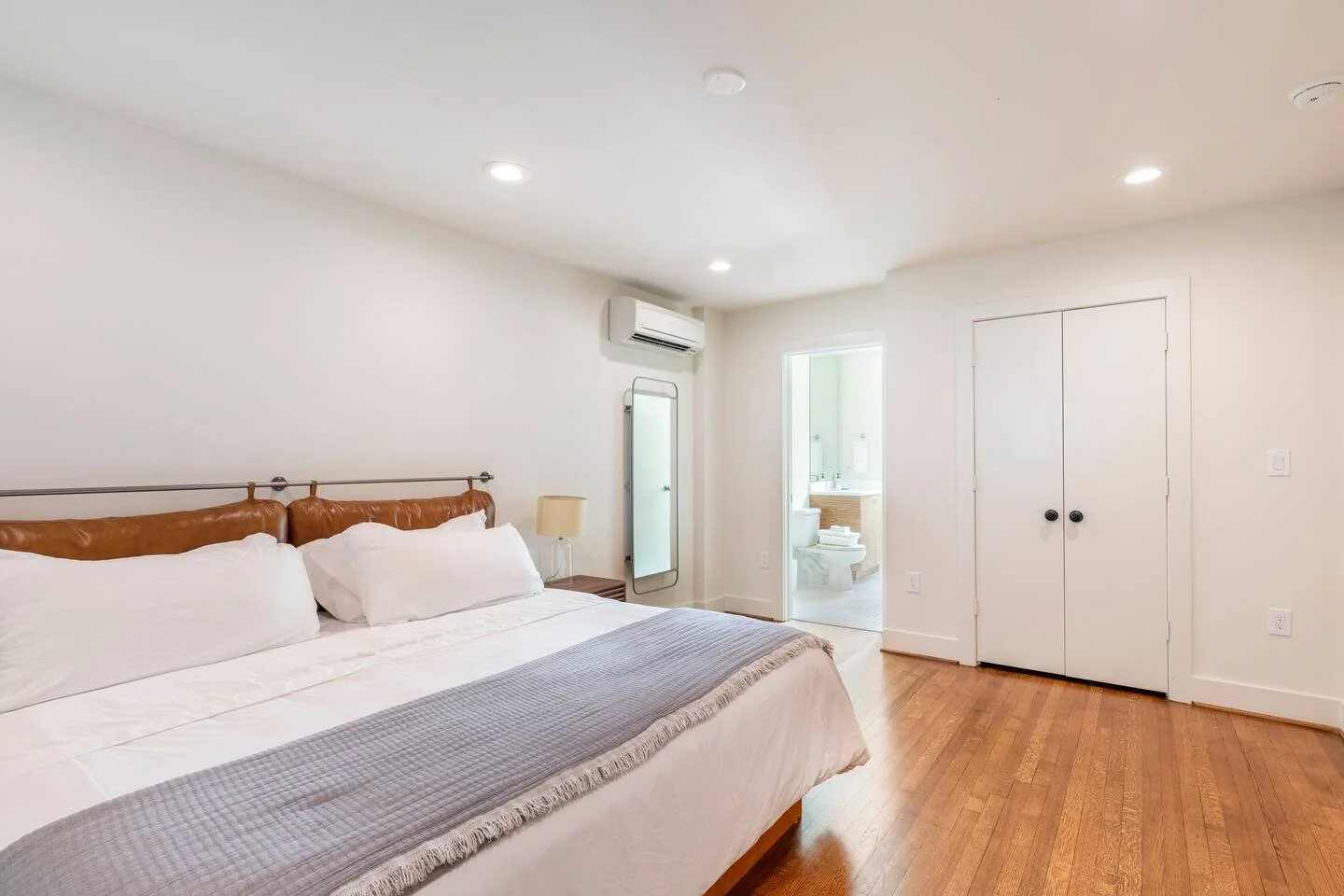 A bedroom with a large bed, white bedding, and a leather headboard. There is a small lamp on a nightstand, a full-length mirror, and an air conditioner. An open door reveals a bathroom with a toilet and sink, and a closet with white doors.