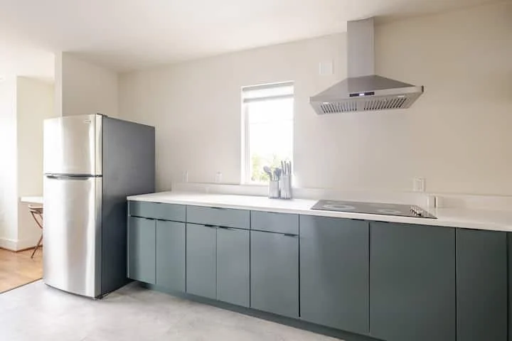 Modern kitchen with blue cabinets, a stainless steel refrigerator, a vent hood, and a small window letting in natural light.