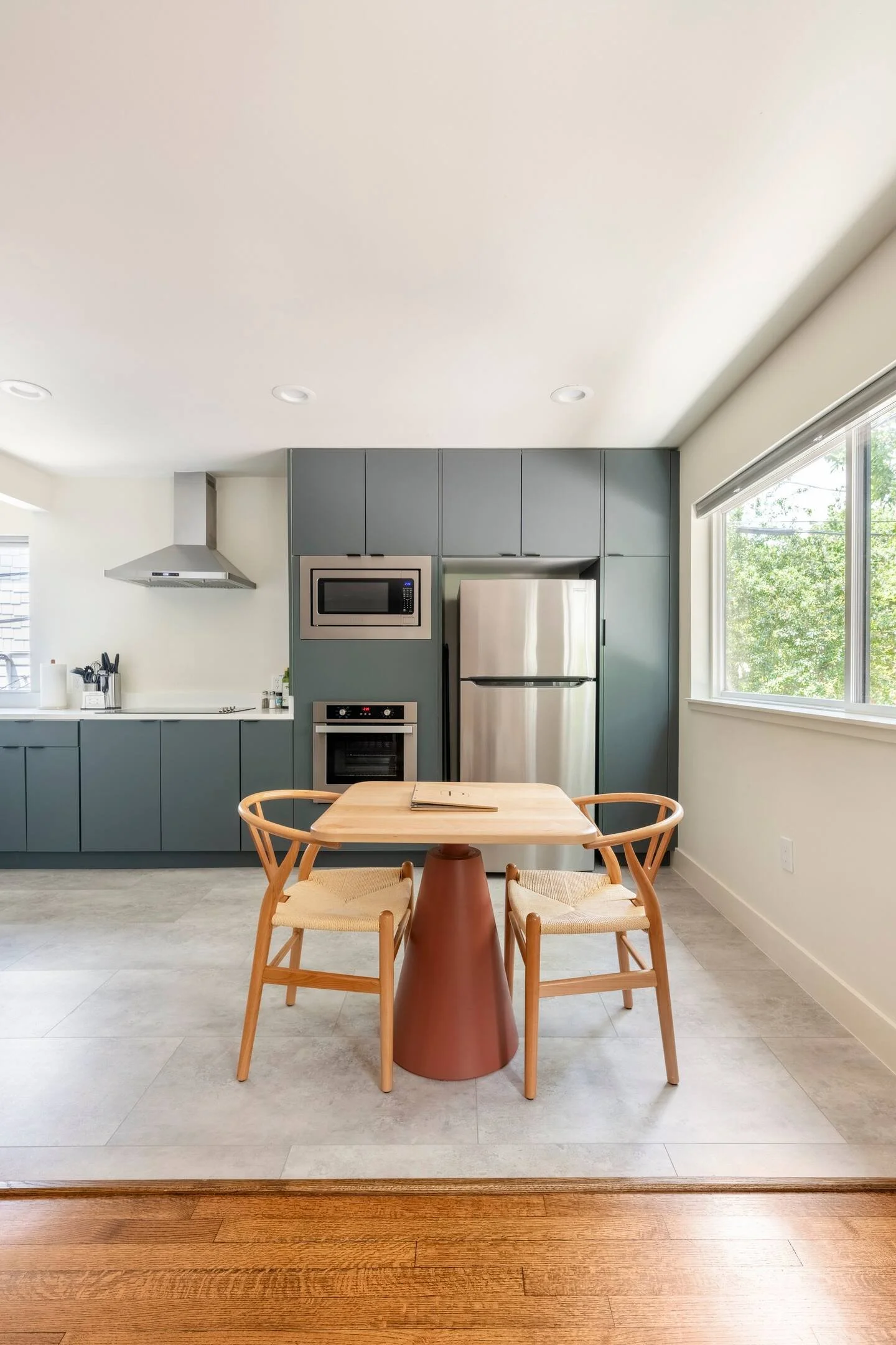 A modern kitchen with gray cabinets, stainless steel refrigerator, microwave, and oven. A small wooden dining table with four matching chairs is in the foreground. Large windows let in natural light and show outside greenery.
