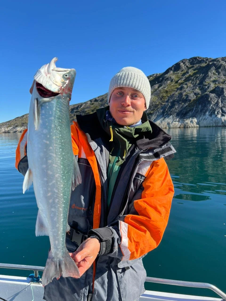 Man i varma kläder som håller i en stor fisk på en båt med vatten och berg i bakgrunden.