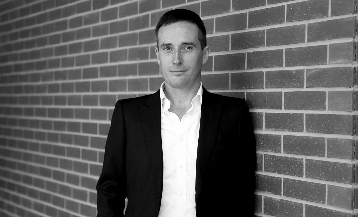 Black and white photo of a man wearing a suit, standing against a brick wall.