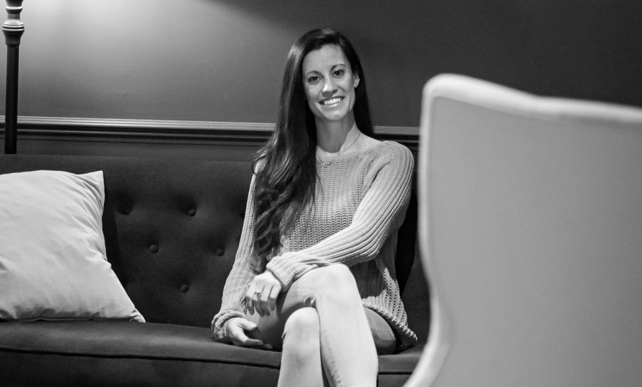 A woman with long, wavy hair sitting on a dark sofa, smiling at the camera, in a room with a pillow and wall-mounted lamp.
