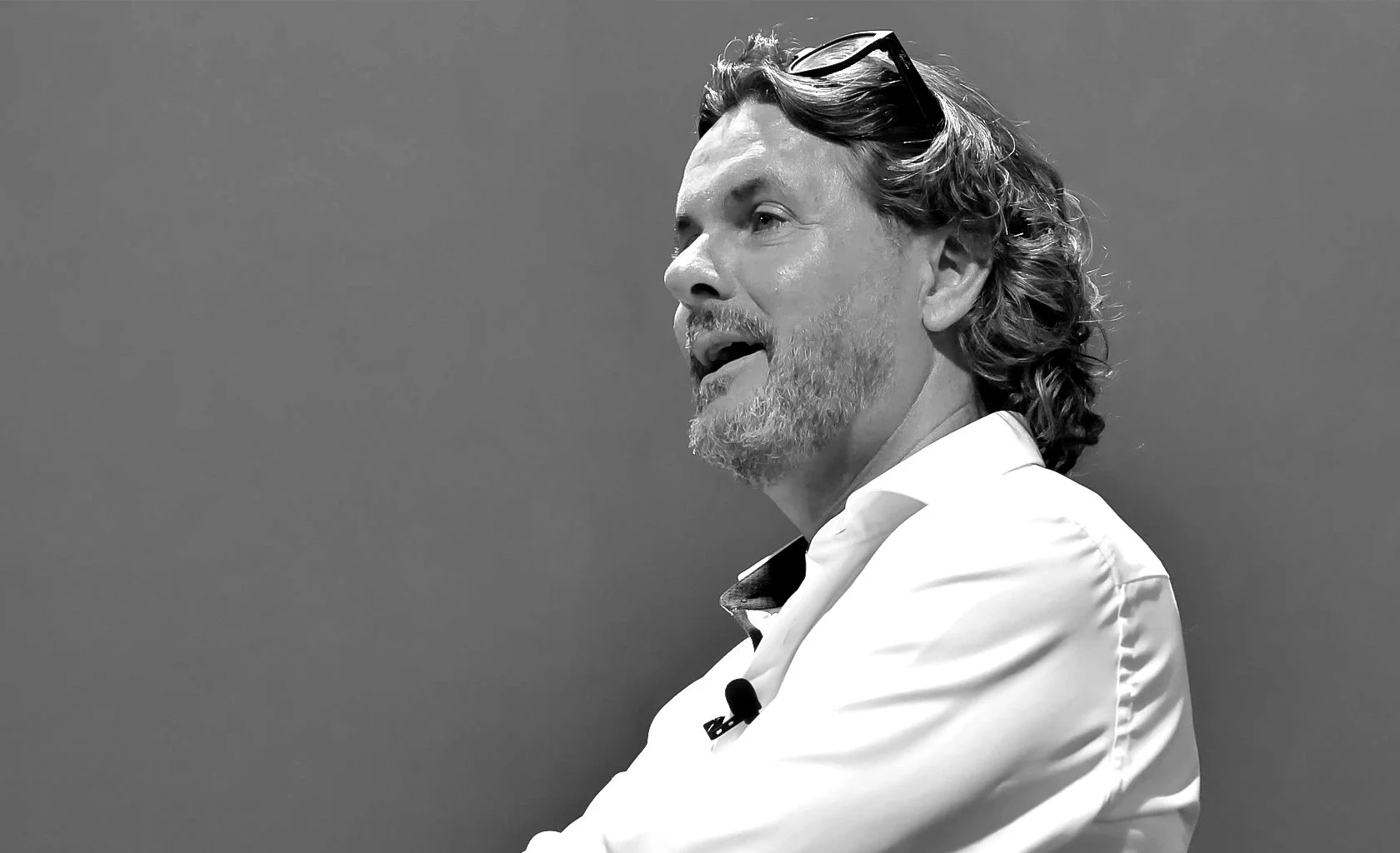 Side profile of a man with wavy hair, facial hair, wearing glasses on his head and a white collared shirt, speaking or explaining against a plain background.