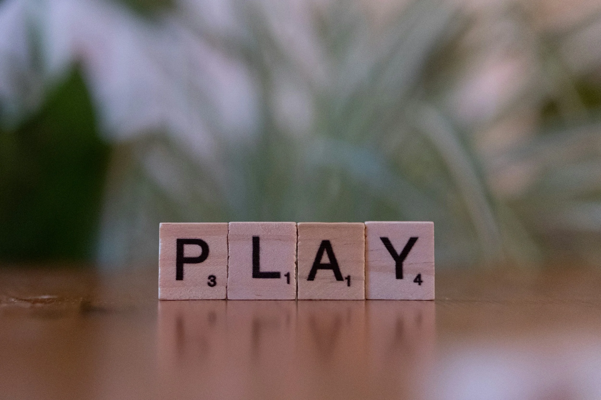 Four wooden scrabble tiles spelling the word "PLAY" on a smooth surface, with a blurred background of green foliage.