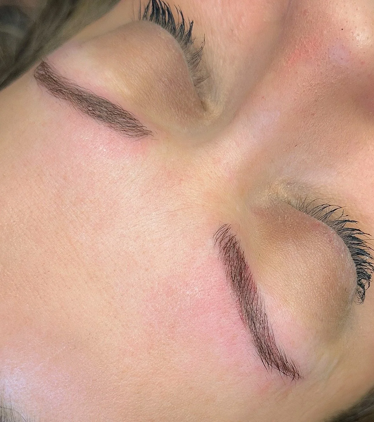 Close-up of a person's face with closed eyes, showing well-groomed eyebrows, long eyelashes, and smooth skin with a slight pinkish hue.