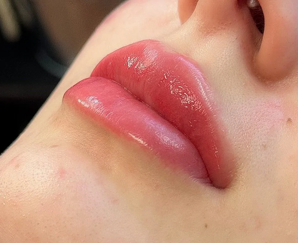 Close-up of a person's lips with glossy pink lipstick, showing a natural skin tone and parts of the nose and cheek.