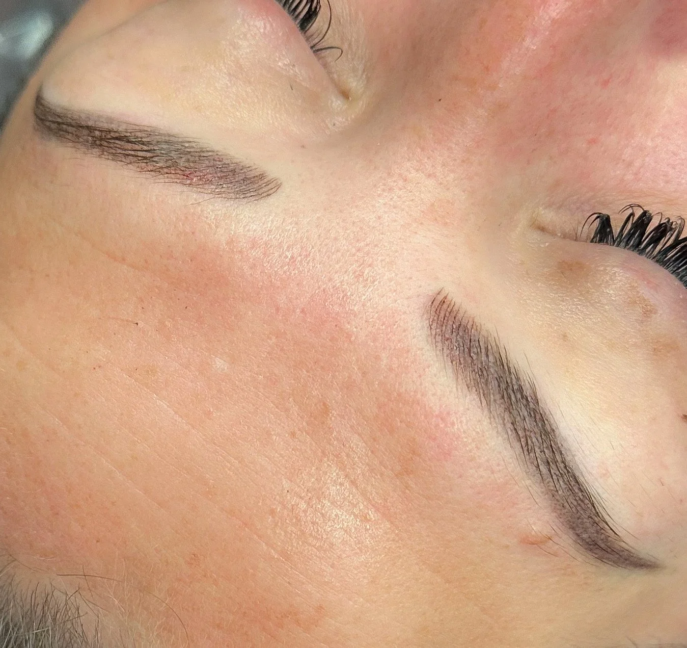Close-up of closed eyes showing freshly shaped and filled eyebrows, long curled eyelashes, and smooth skin with slight redness.