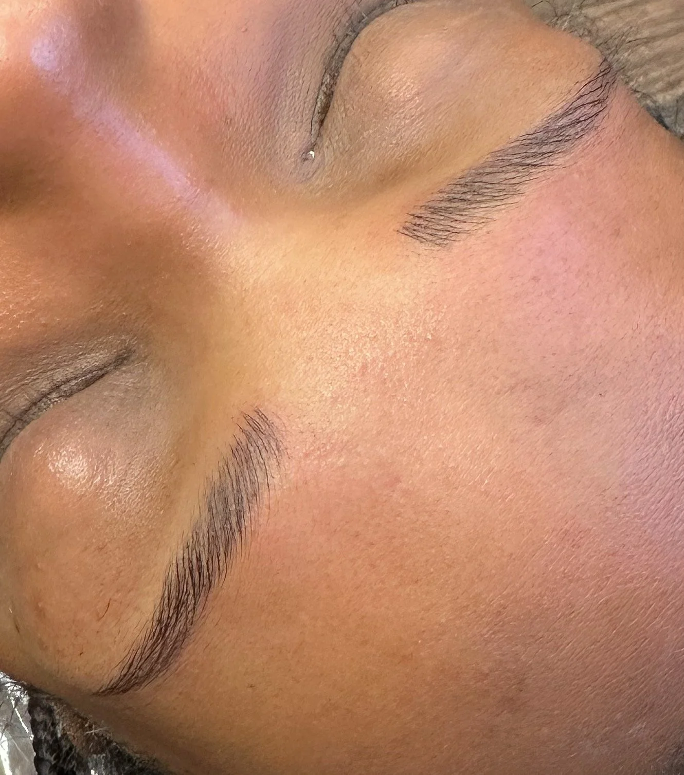 Close-up of a person's face with closed eyes, showing well-groomed eyebrows, clear skin, and a small nose piercing.