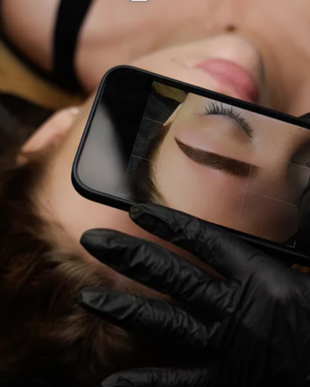 A person with brown hair lying down receives a close-up eye makeup consultation, with a mirror reflecting their eye with winged eyeliner.