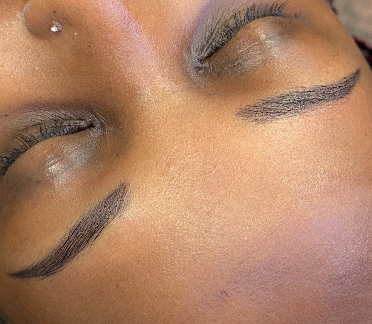 Close-up of a person's face with closed eyes, showing detailed eyebrow makeup, long eyelashes, smooth skin, and a small nose piercing.