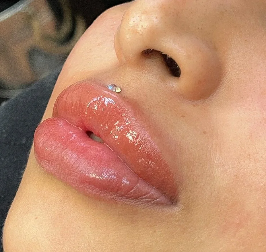 Close-up of a person's lips with glossy, nude lipstick, a small piercing above the upper lip, and part of their nose visible.