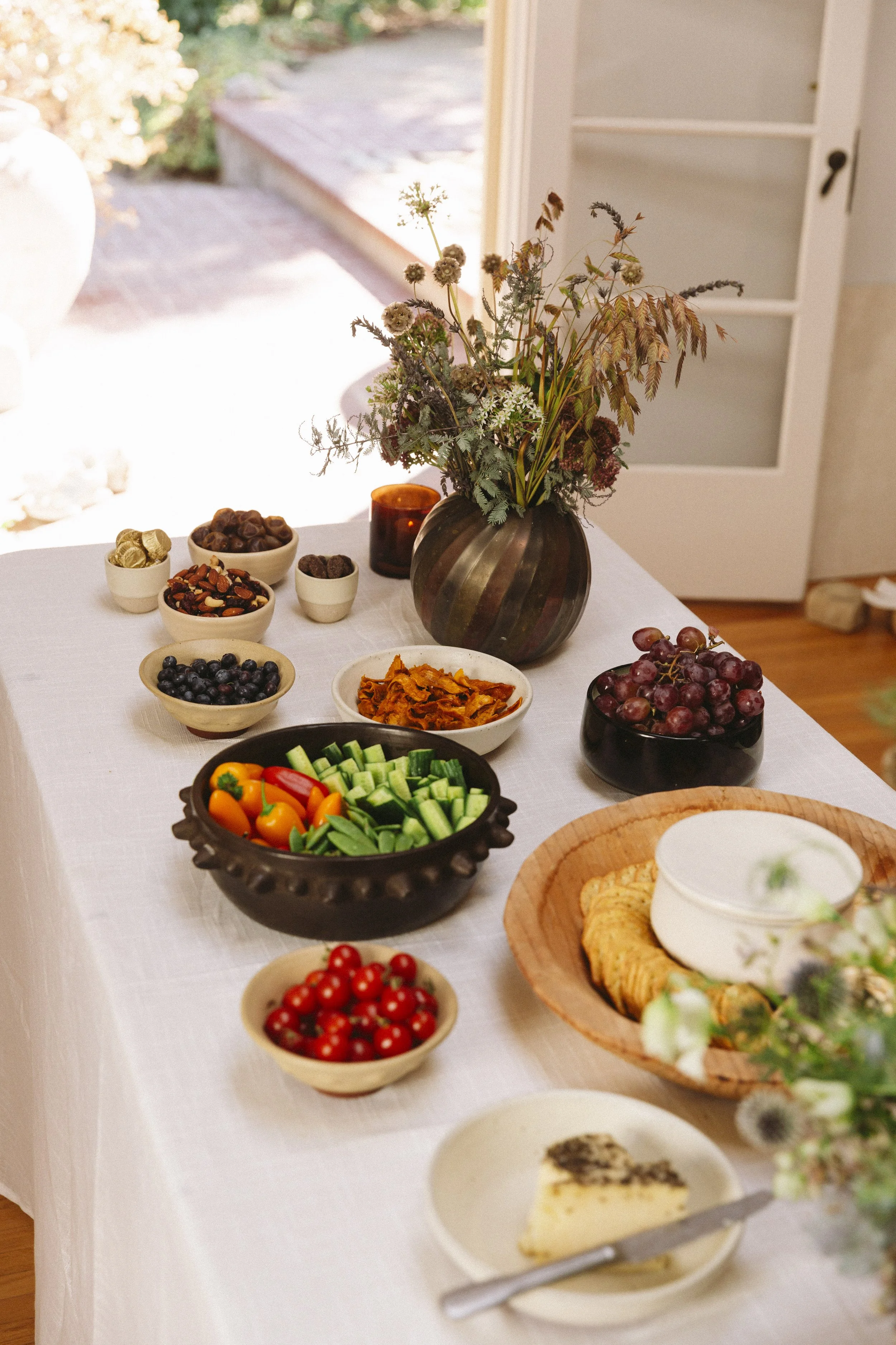 A table set with a variety of bowls filled with colorful fruits, vegetables, nuts, and a cheese platter, with a vase of wildflowers in the background in a bright room.