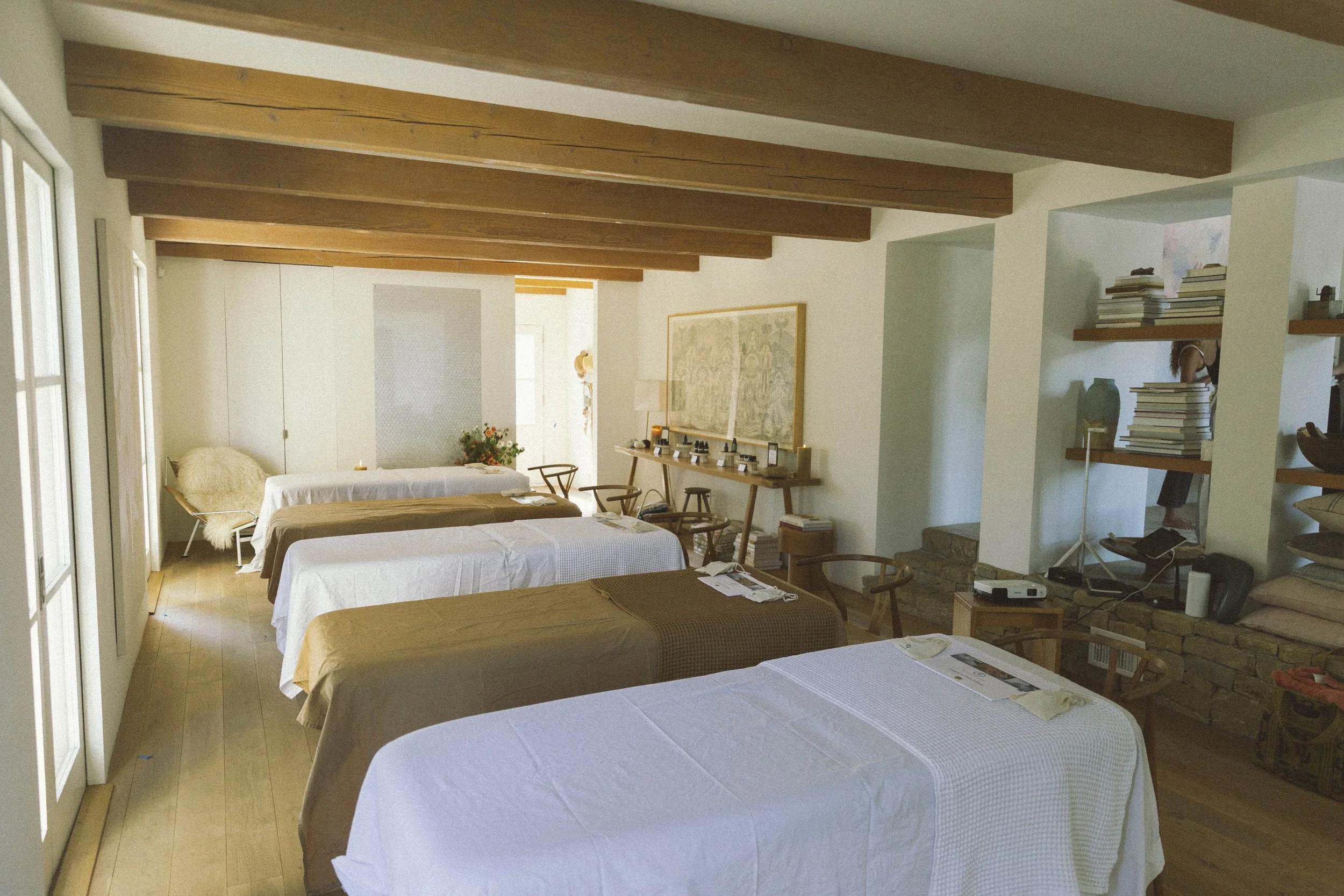 A room with five beds covered with white and brown sheets, wooden floors, ceiling beams, and bookshelves on the right side.