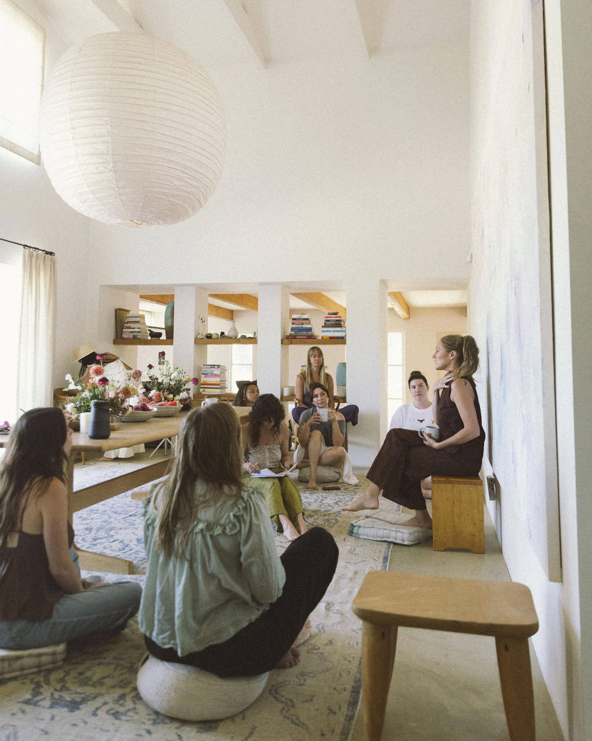 Group of women participating in a discussion in a bright, modern living room with white walls, a large paper lantern, and bookshelves.