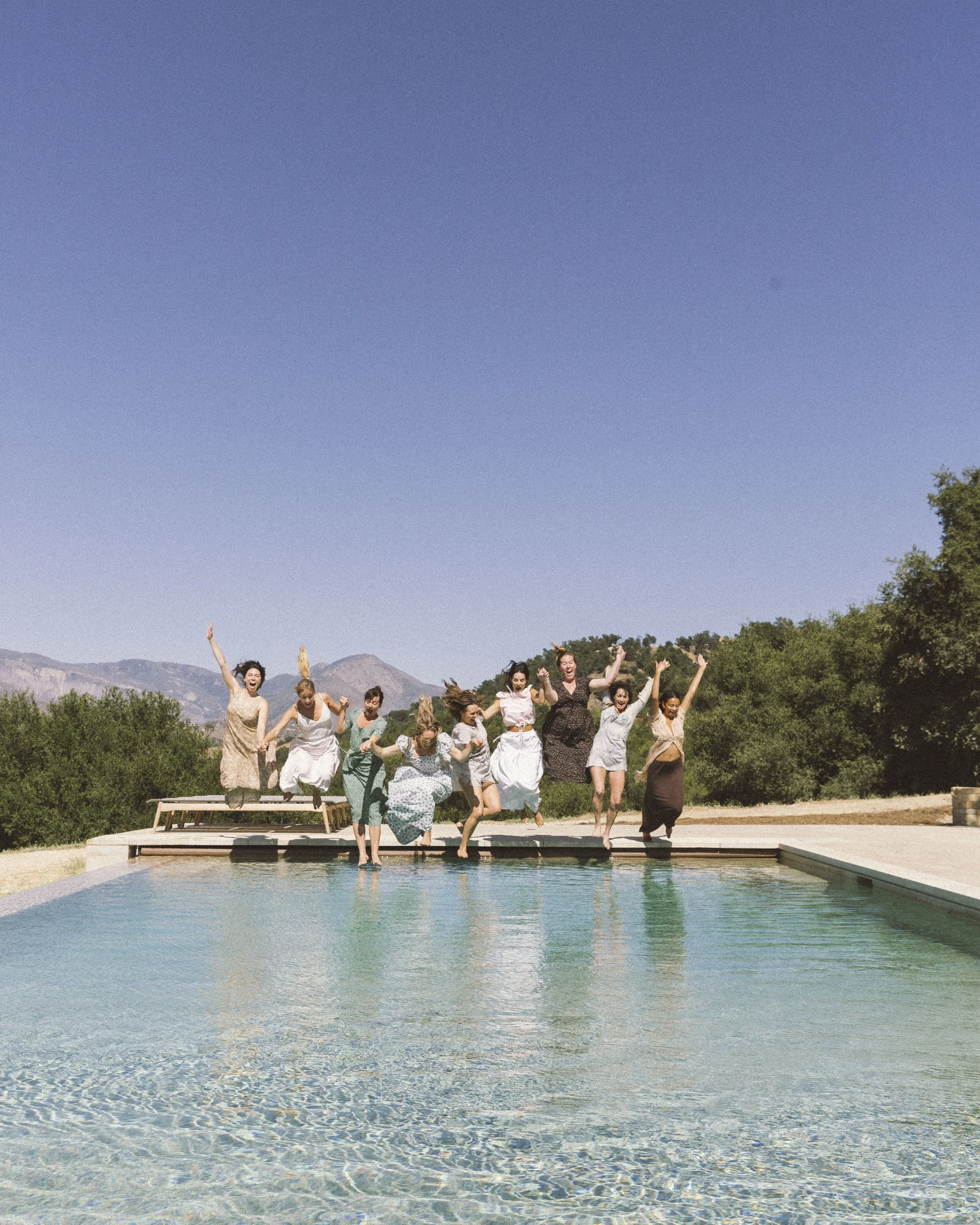 Group of women in vintage dresses jumping from a dock into a pool on a sunny day, with mountains and trees in the background.