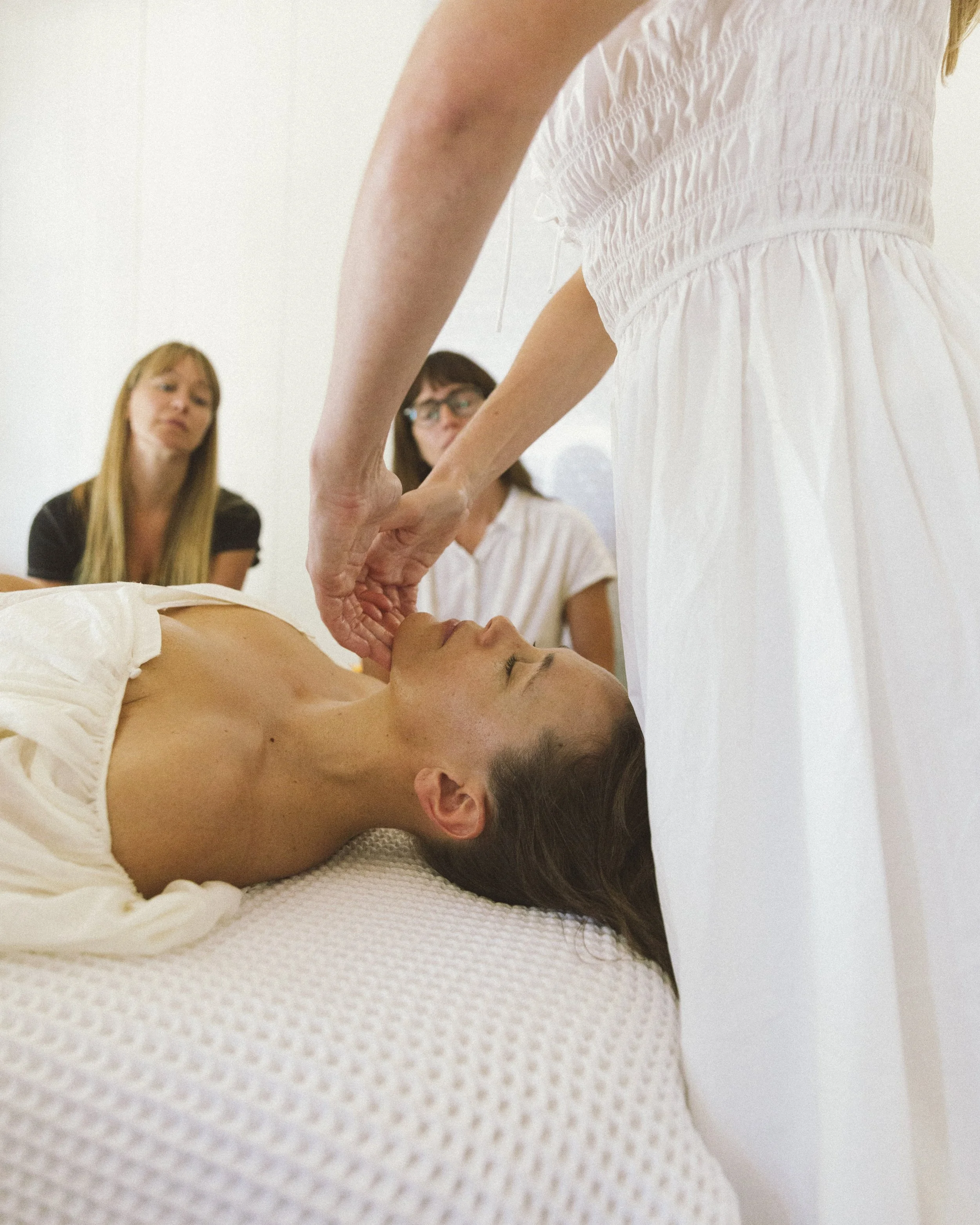 A woman receiving a facial treatment while lying down on a spa bed with her eyes closed, with two women observing in the background.