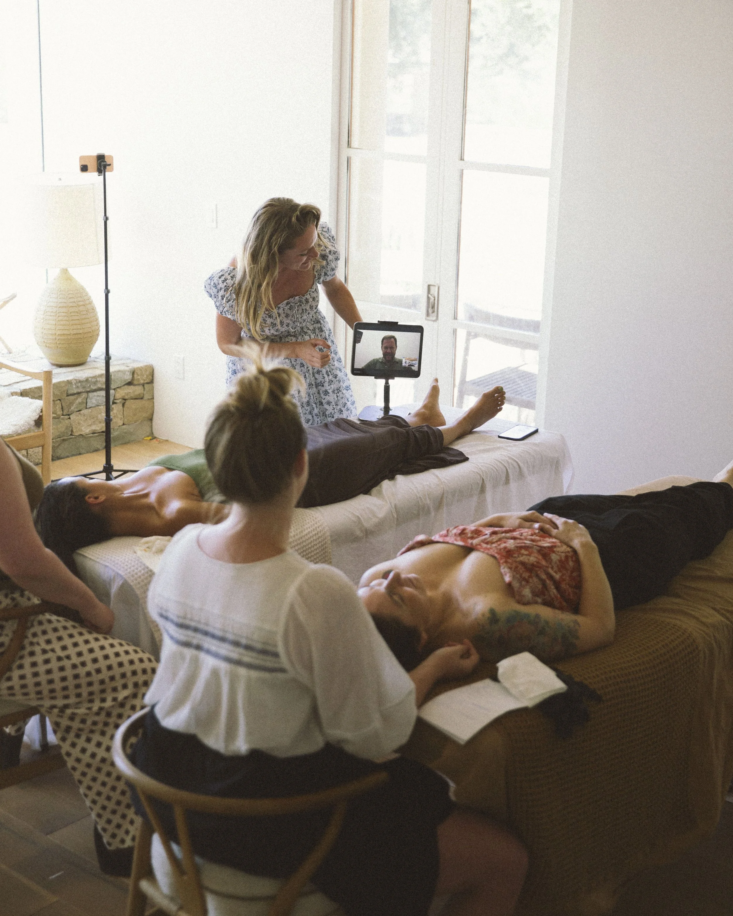 Three women rest on massage beds, one woman lies face down, another woman lies on her back, and the third woman is seated, taking notes or writing.
