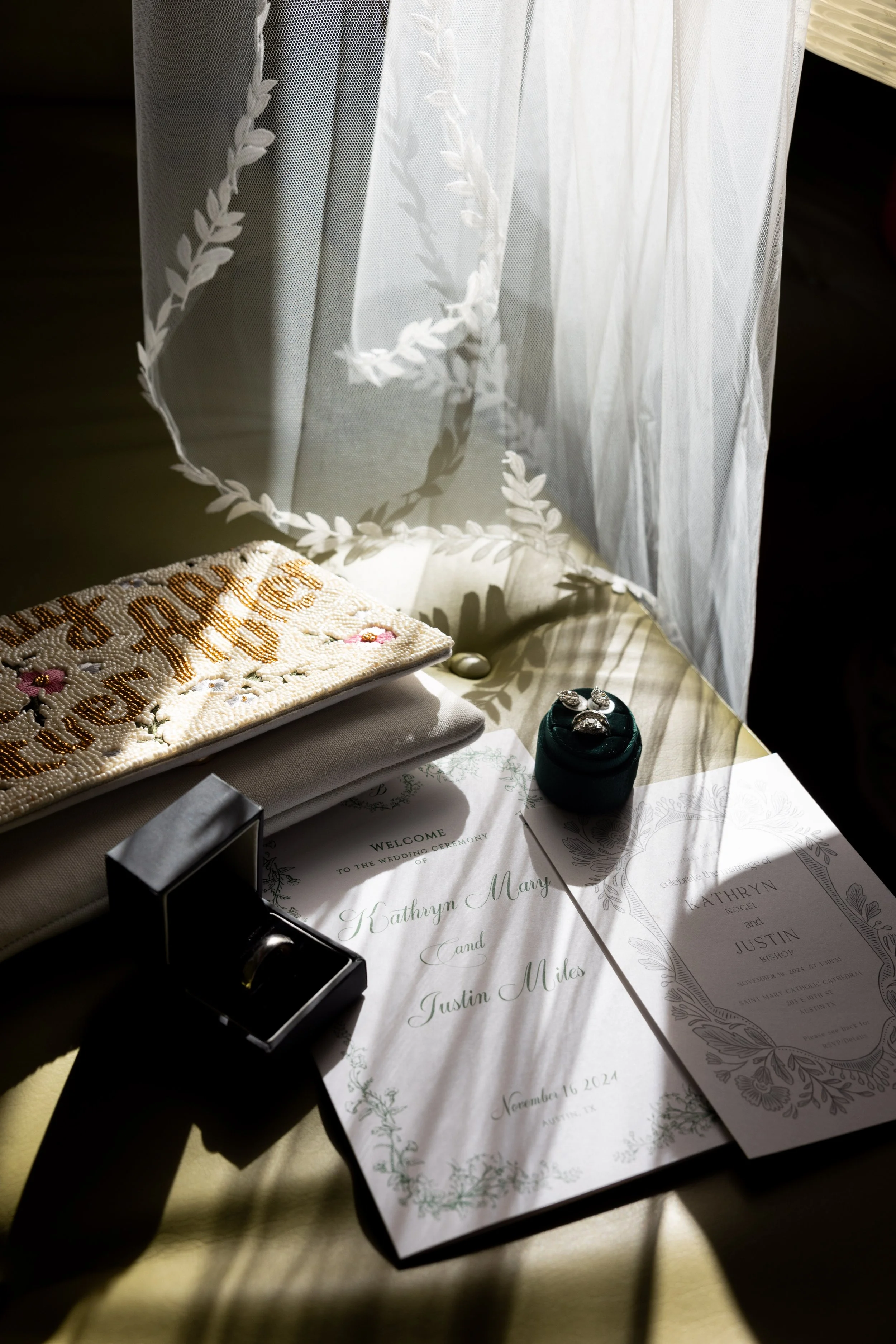 Wedding invitation, rings, and jewelry displayed on a table with dappled sunlight and sheer curtains in the background.