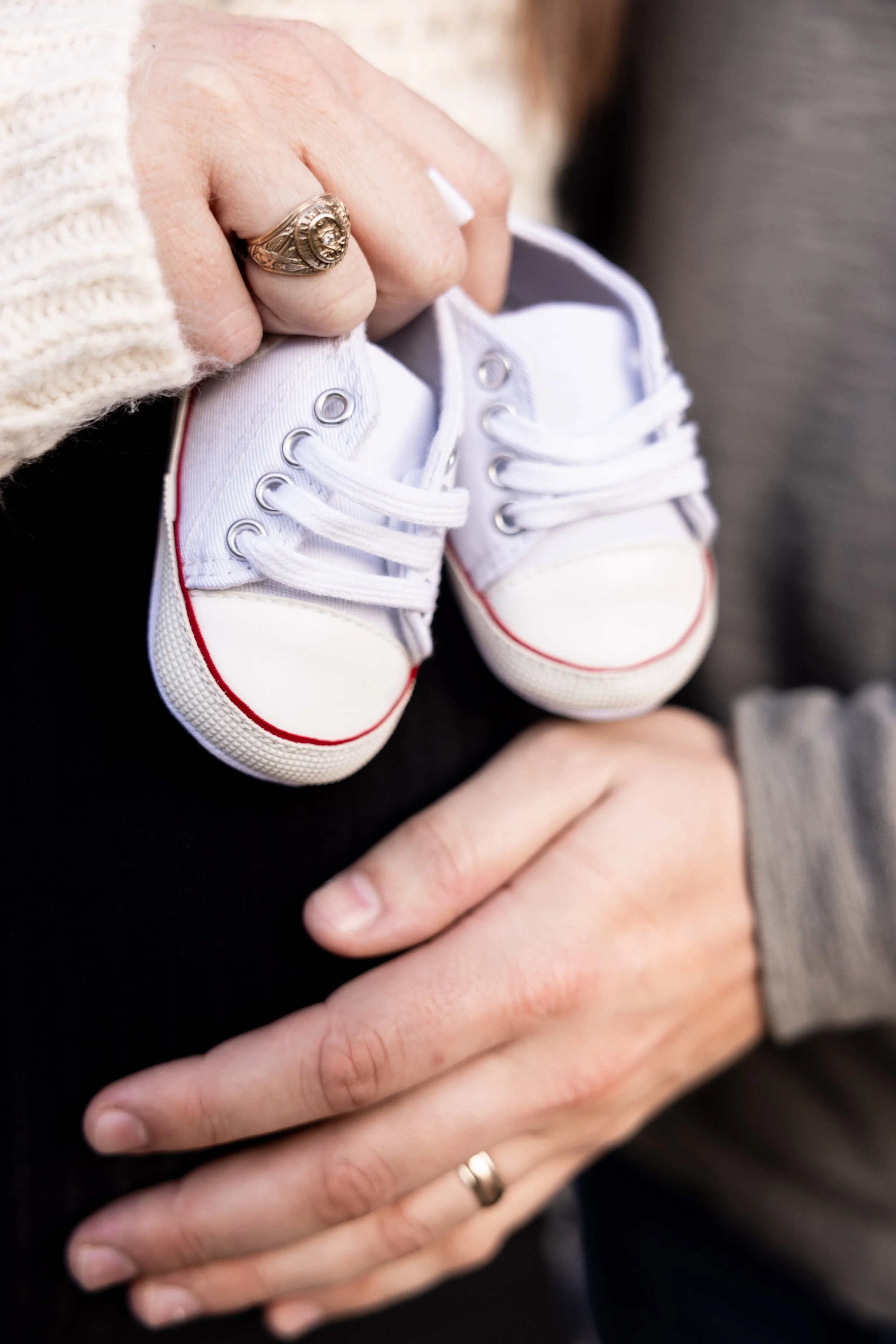 A pregnant mother holding a pair of tiny white sneakers with red trim, wearing a beige sweater and a wedding ring.