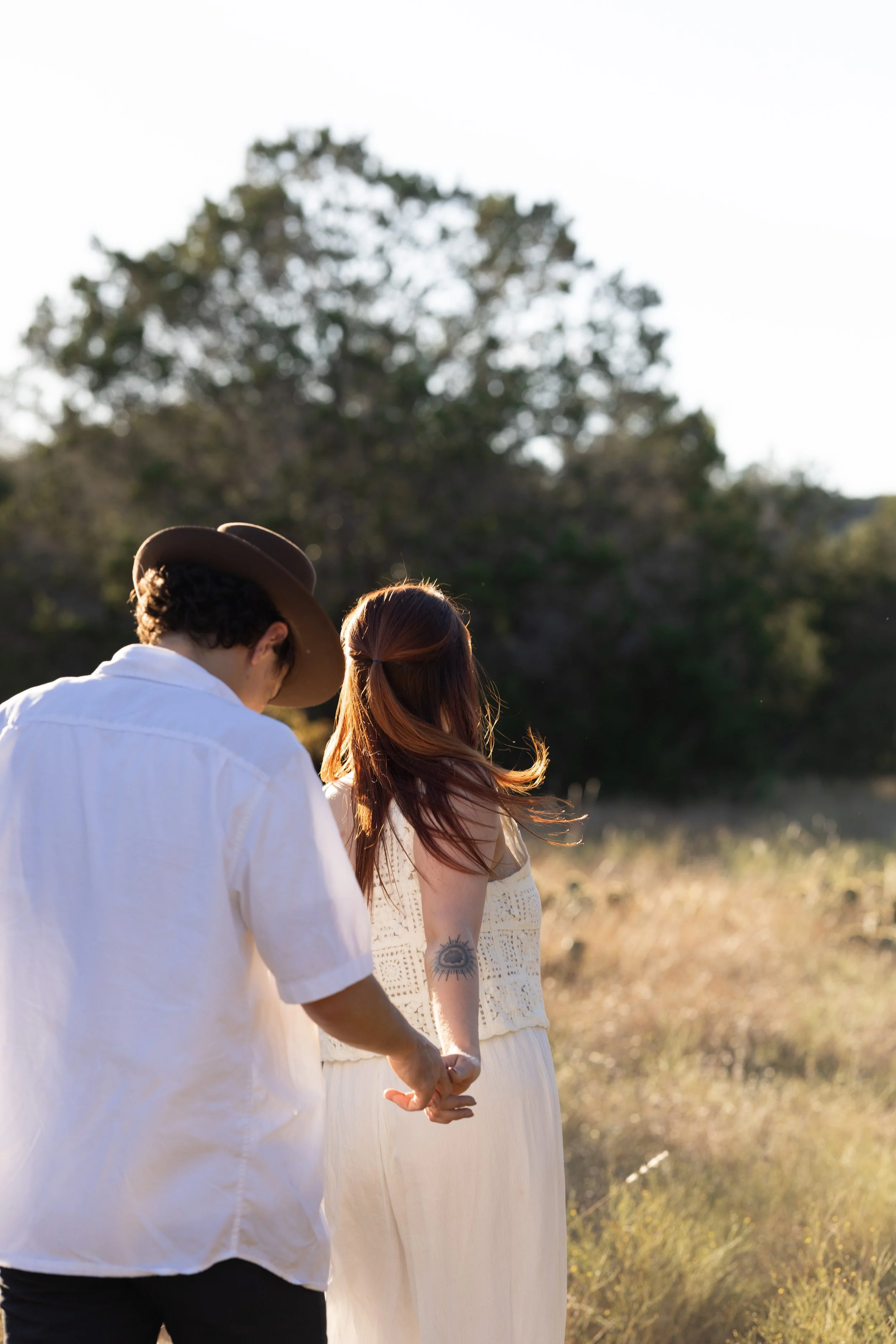 An engaged couple walking hand in hand through a grassy field with trees in the background during sunset.