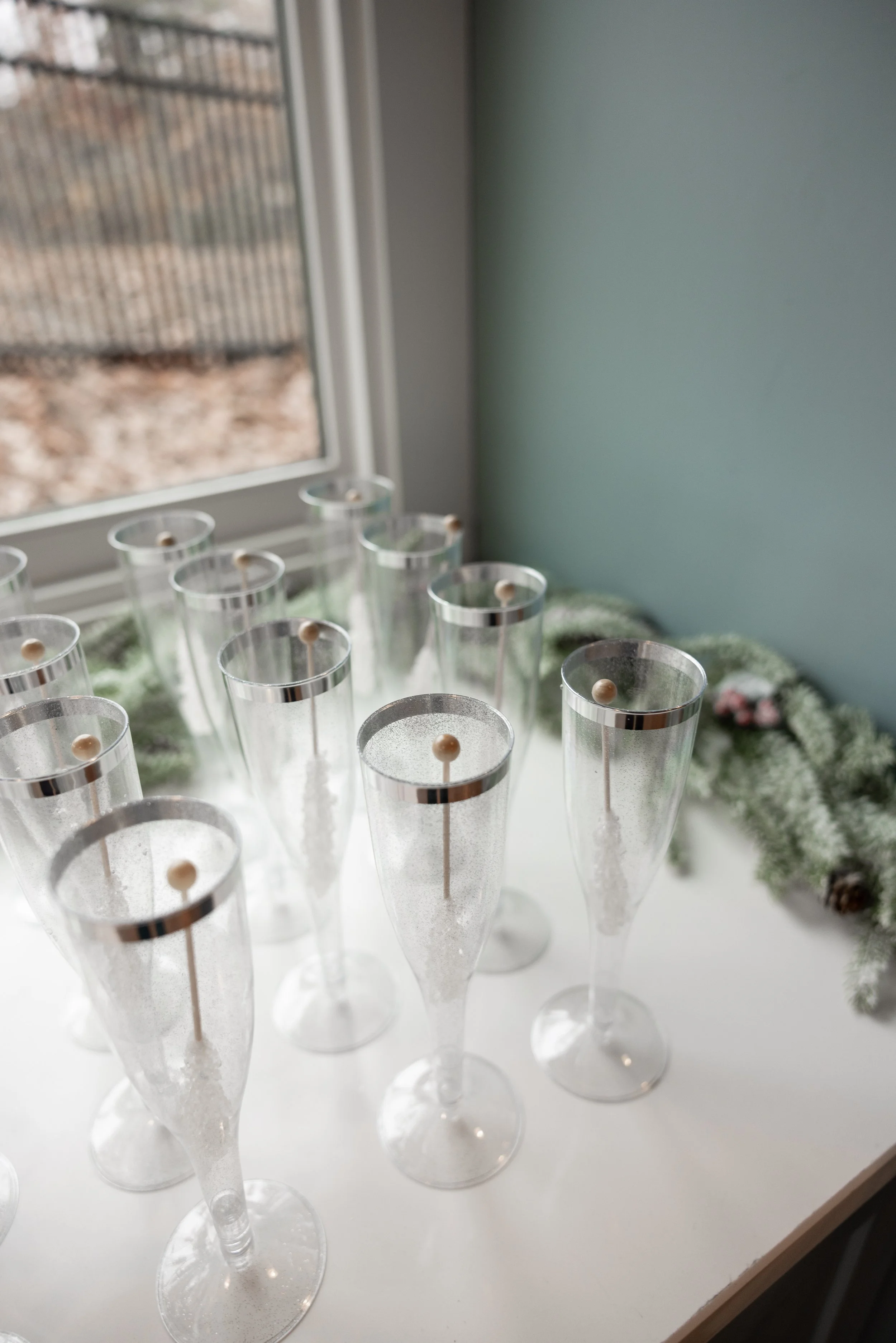 Several empty champagne flutes with silver rims and wooden stir sticks inside, arranged on a white surface near a window, with a Christmas greenery decoration in the background.