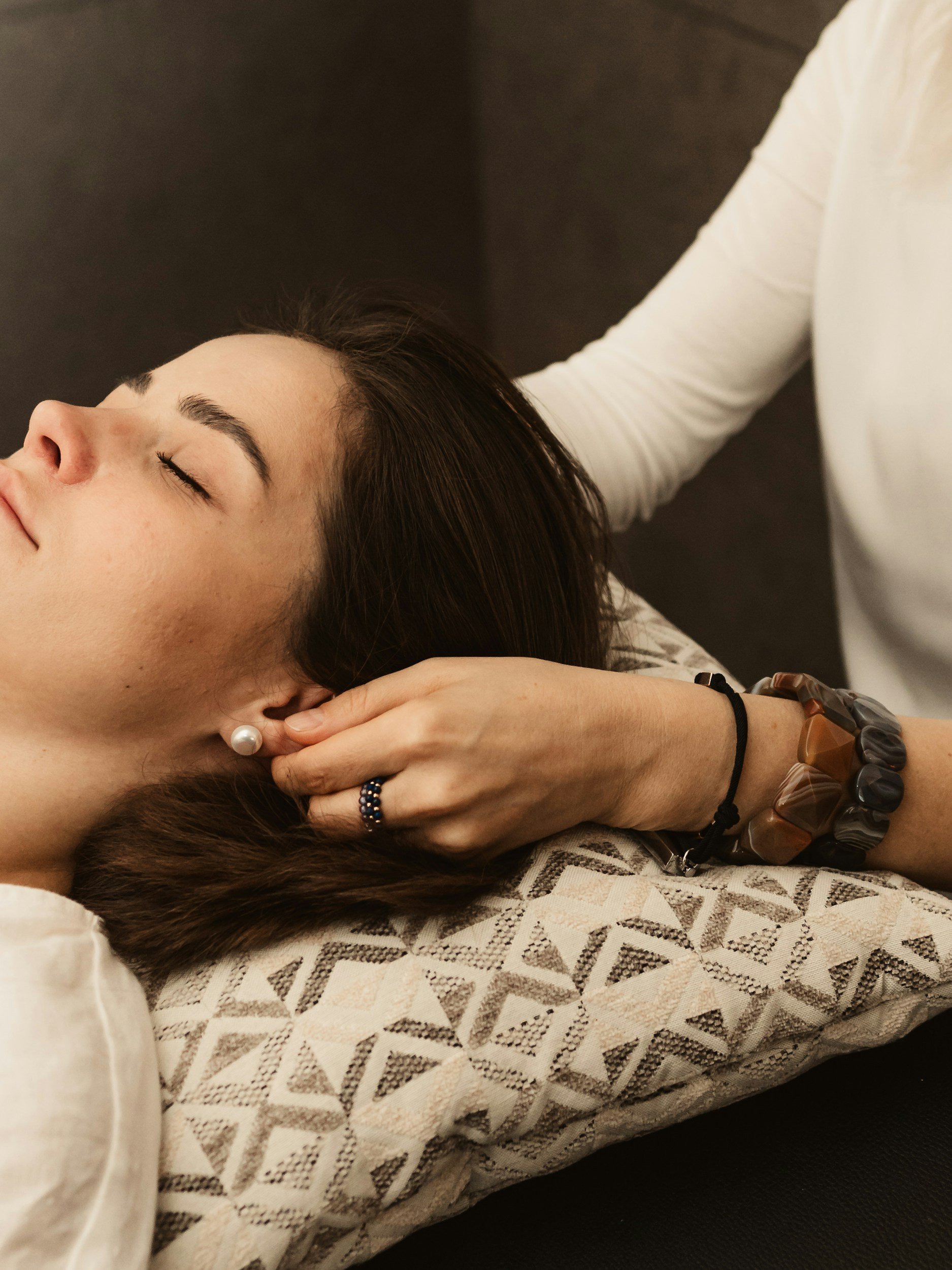 A woman is lying down with her eyes closed, receiving a massage or therapy on her head and neck from another person. The woman has dark hair, pearl earrings, and a ring on her finger. She is resting on a patterned cushion.