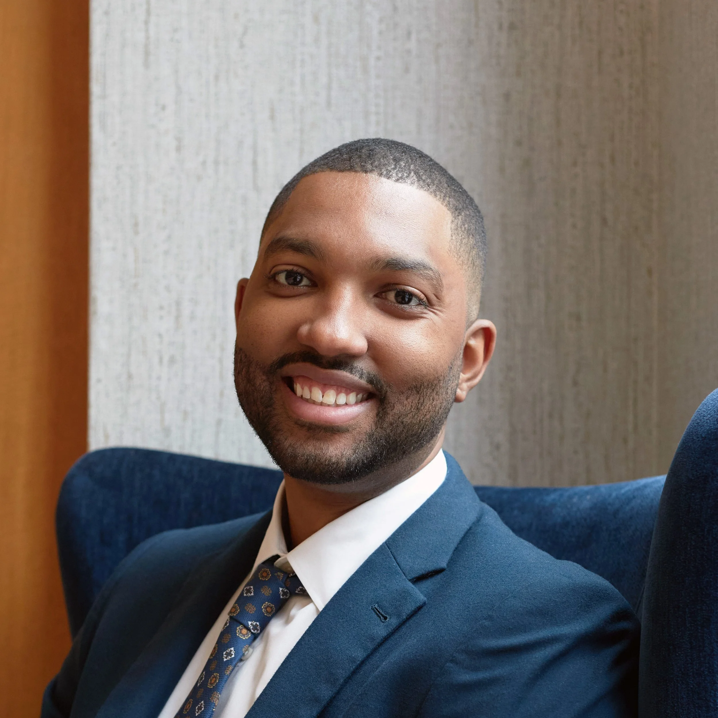 A young man with short hair and a beard smiling, wearing a navy blue suit, white shirt, and patterned tie while sitting on a blue chair with a textured wall background.