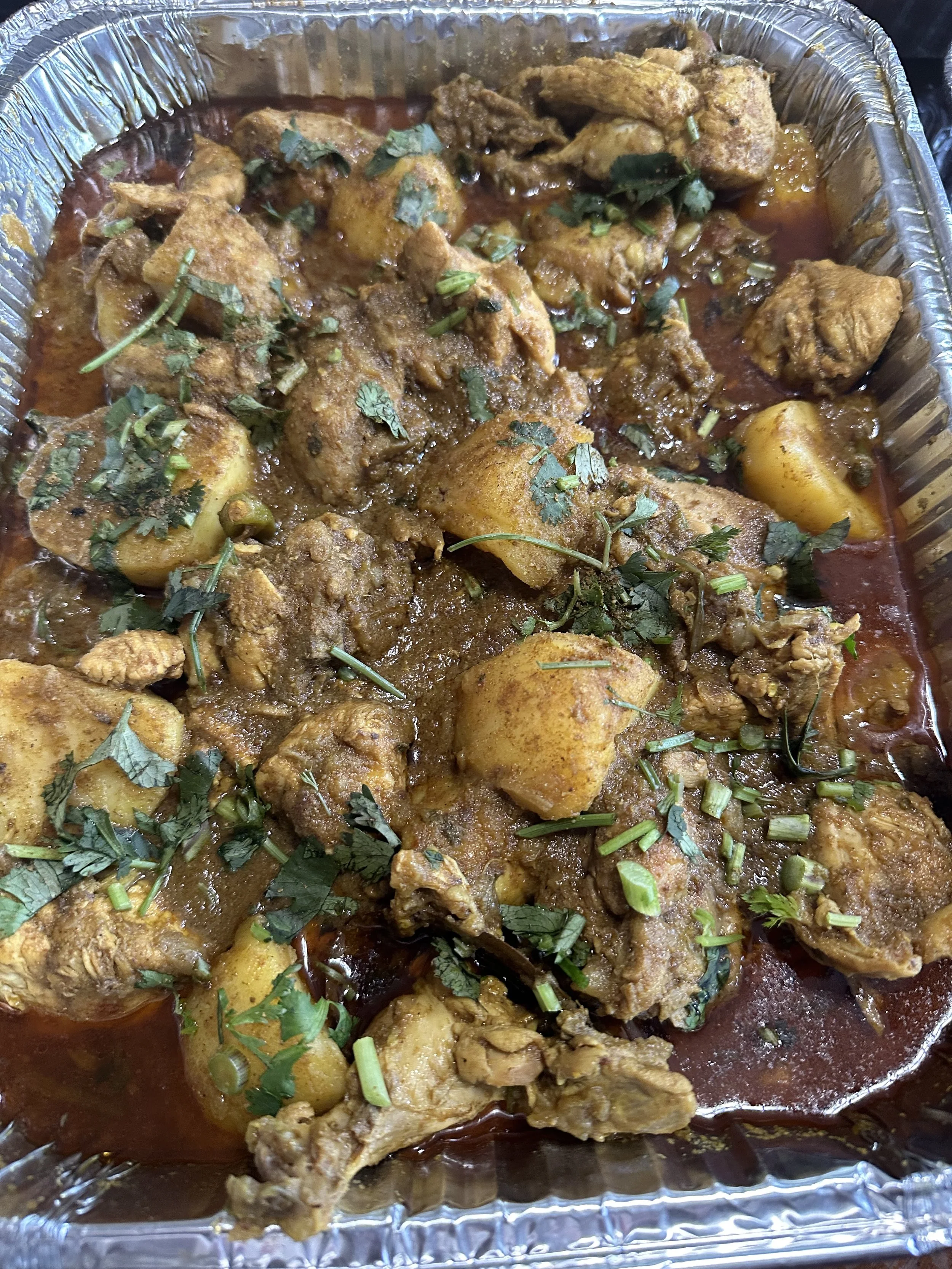 Bengali chicken curry with potatoes in a rich brown gravy, garnished with cilantro in a foil catering tray.