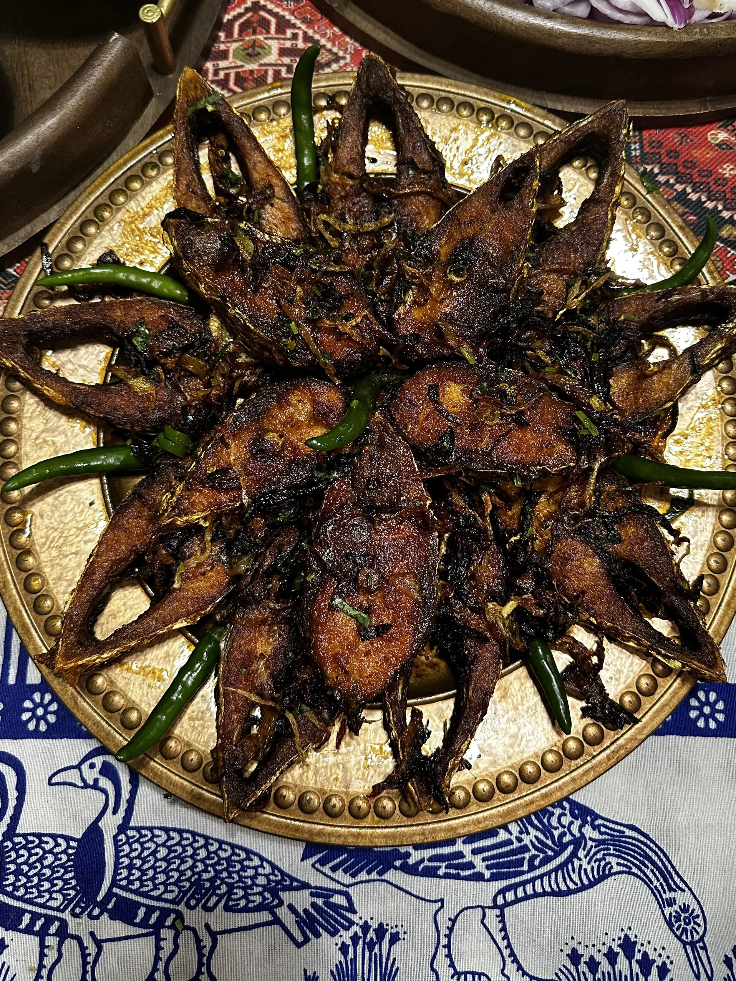 Fried fish steaks topped with browned onions and green chilies, arranged on a decorative gold platter.