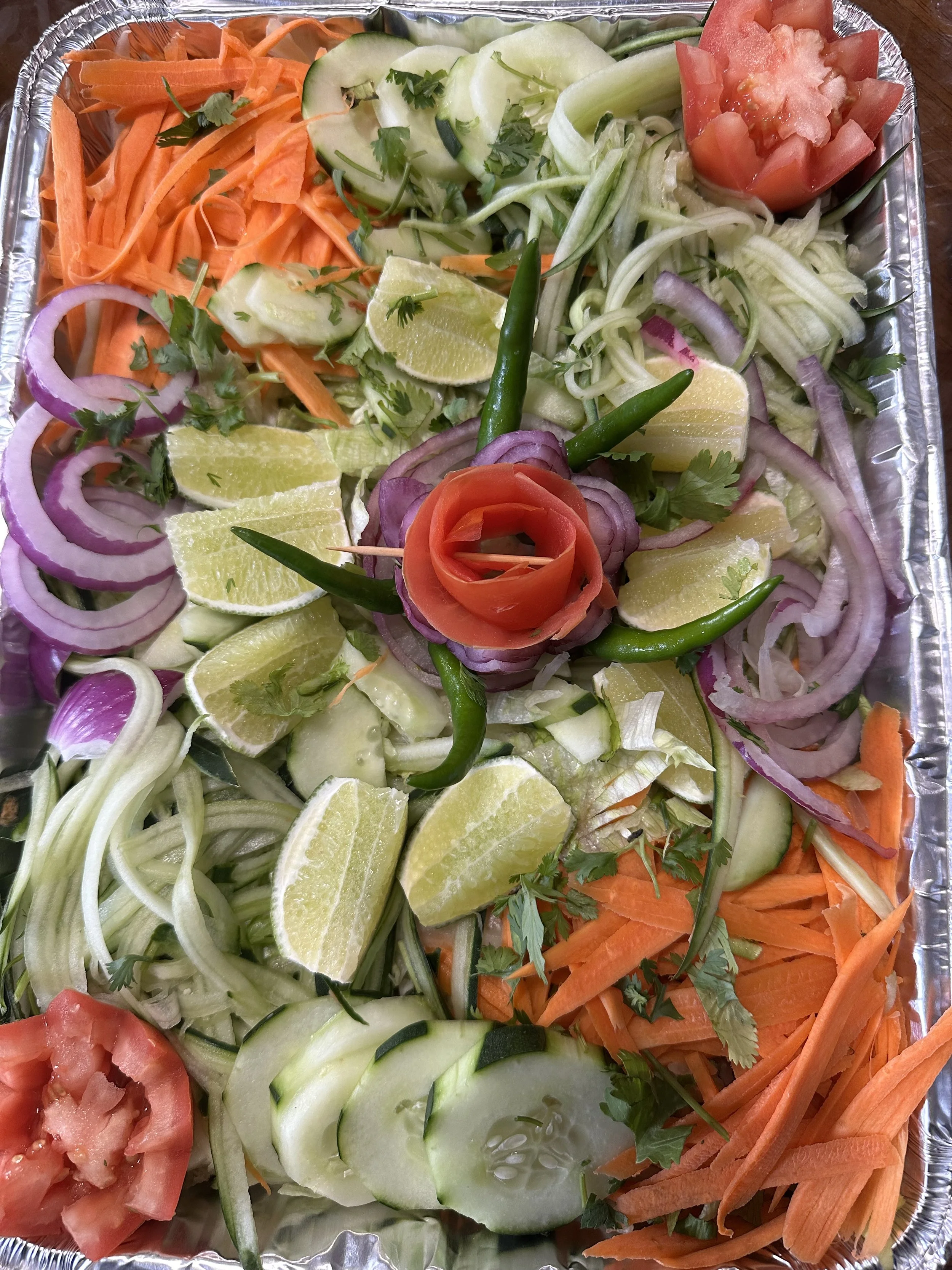 Catering tray of cucumber, carrot ribbons, red onion, lime wedges, and green chilies, decorated with carved tomato flowers.